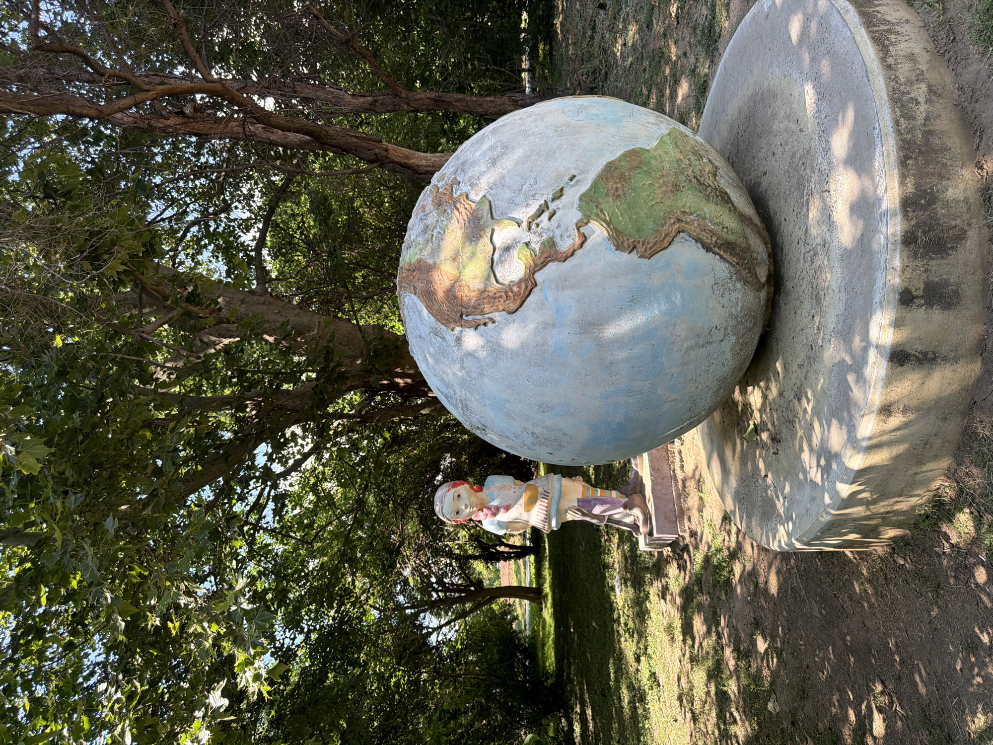 In Pazardzhik, Bulgaria, a statue of a young girl stands in a park, looking at a large, weathered globe. The girl, with braided red hair and a blue and white dress, holds a yellow bowl in her hands. The globe, with its continents painted in brown and green, sits on a concrete pedestal. The scene is set under the shade of large trees, with dappled sunlight falling on the grass and dirt ground. The overall impression is one of a quiet, outdoor art installation.