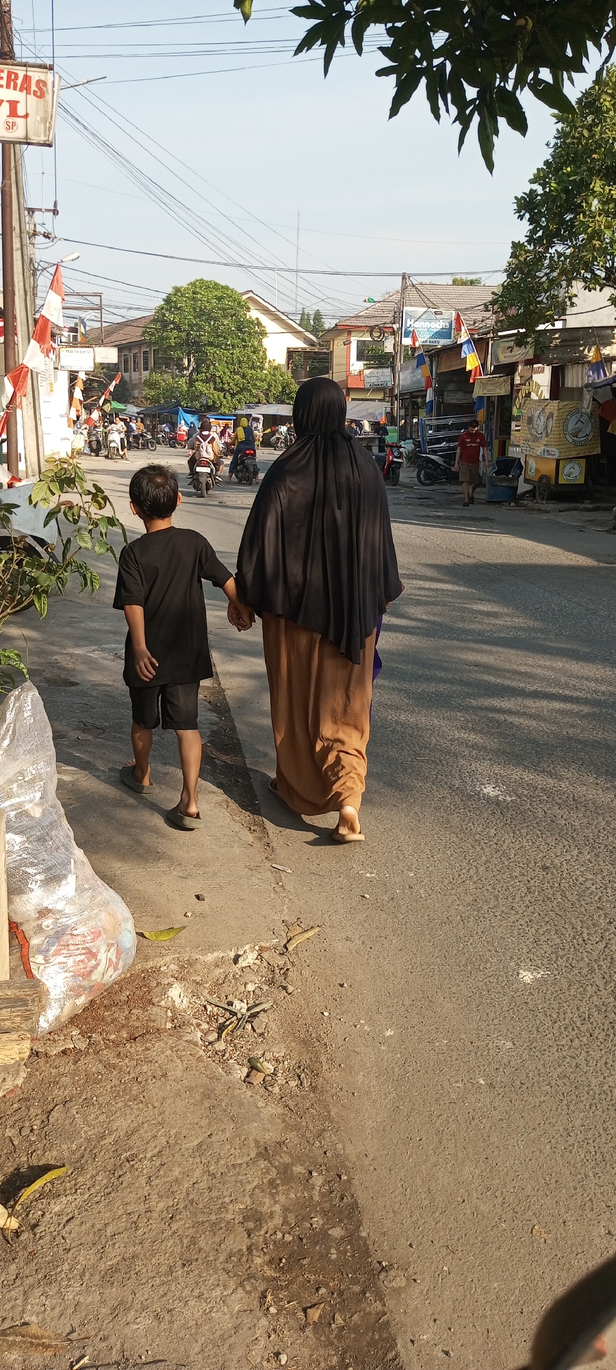 In Setu, Indonesia, on a paved street under a clear sky with dappled sunlight, a person wearing a black headscarf and a long, tan skirt walks hand-in-hand with a young boy in a black t-shirt and shorts. They are walking away from the camera, towards a street lined with buildings and shops. Several motorcycles are visible in the background, some parked and some in motion, suggesting moderate traffic. A plastic bag filled with items sits on the edge of the sidewalk to the left of the frame, next to some plants and debris. The overall scene depicts a daily street view with pedestrian activity and local commerce.