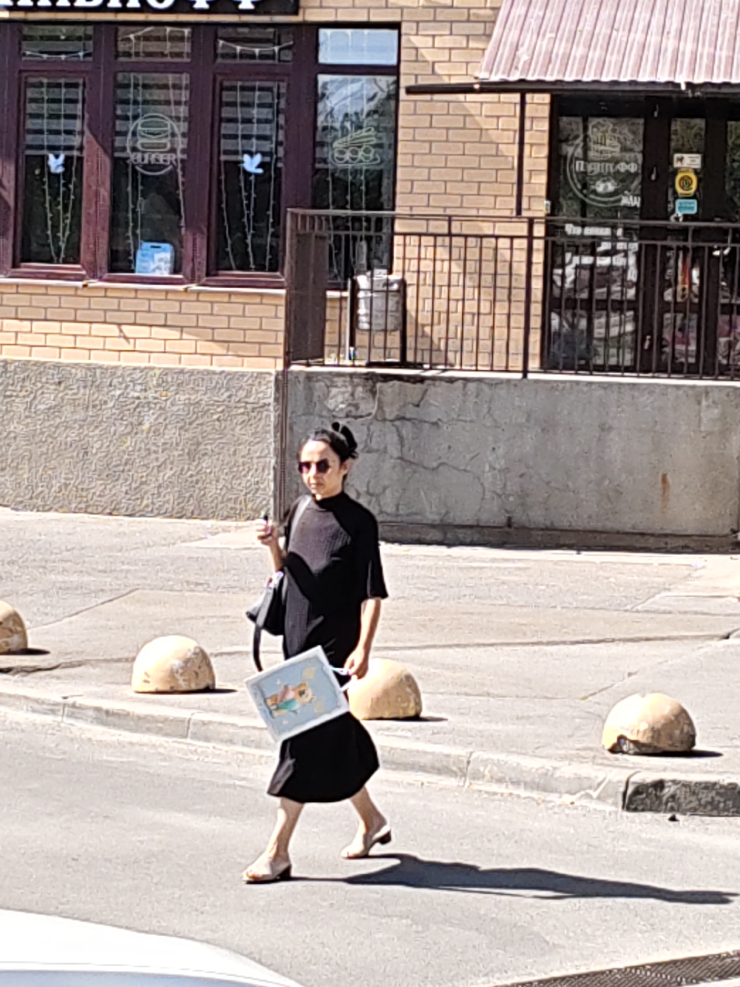 A woman in a black dress and sunglasses walks across a street in Rostov-on-Don, Russia, holding a picture. She has her hair in a bun and is carrying a black purse. The background shows a building with windows and a metal fence. There are also several rounded concrete bollards on the side of the road. The lighting suggests it is daytime and sunny.