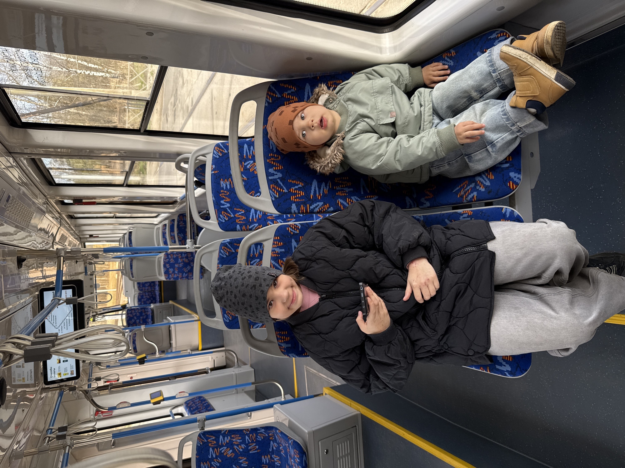 Two children are lying on seats on a train. The boy wears a brown patterned beanie, a light green coat with fur around the hood, blue jeans, and brown boots. The girl wears a gray patterned beanie, a black puffer jacket, and gray sweatpants. The seats are blue with a geometric pattern. The train has a skylight. The train car has route information displayed on an electronic device, and is in Rusanovo, Russia.