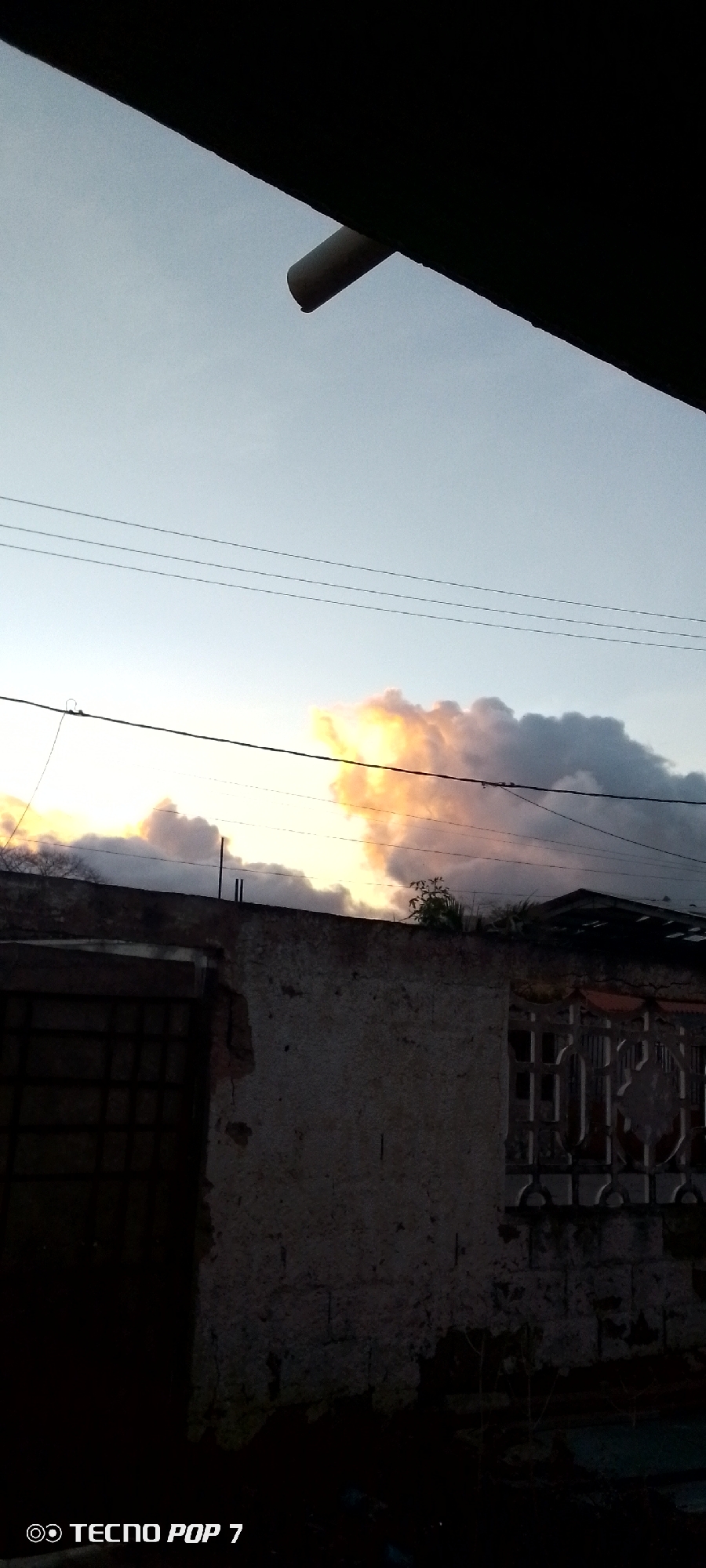 The image captures a scene during either sunrise or sunset, characterized by a dramatic sky over what appears to be an urban or semi-urban setting in Altagracia, Venezuela.
The upper portion of the frame features a clear, light blue sky transitioning into a vibrant display of clouds illuminated by warm, golden, and orange light, contrasting with darker grey cloud masses. Three thin power lines stretch horizontally across the upper sky, with a thicker black cable positioned lower down, partially obscuring the brightest part of the clouds.
In the foreground, a dark, undefined structure or roof overhang angles down from the top left, from which a white, cylindrical drain pipe extends towards the center. Below the sky and power lines, the scene is dominated by the upper part of a weathered, textured wall, possibly made of concrete or stucco, showing signs of age and slight decay. Rebar or thin metal rods protrude from the top of this wall. On the left, a section of a dark, barred window or gate is visible, while to the right, a more ornate, patterned metal gate or fence can be seen, partially obscured by the wall. Sparse, small green vegetation sprouts from cracks or atop the wall and nearby structures. The overall foreground is dark, silhouetted against the bright sky, enhancing the twilight effect.
No people or explicit activities are visible. The only discernible text, "TECNO POP 7," is located in the bottom left corner, indicating the device used to capture the photograph.