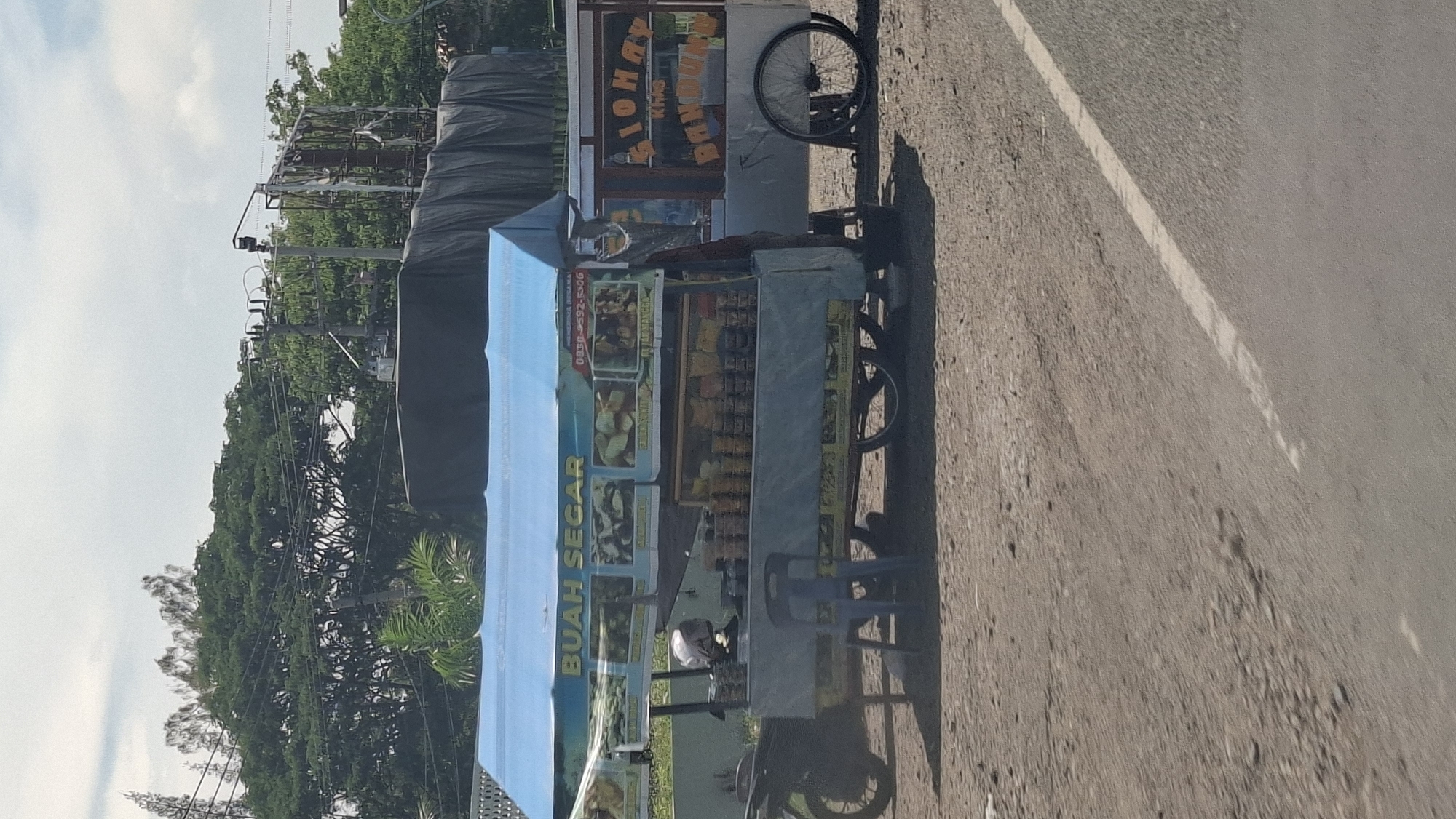 In Cotbadatunong, Indonesia, a roadside fruit cart with a blue awning displays various fresh fruits. The cart is parked on a dirt road next to a paved road with a white line. Behind the cart, a motorcycle is parked, and a blue plastic chair sits in front of it. The cart's display is covered with images of fruits and text, including "BUAH SEGAR" which translates to "FRESH FRUIT," and a phone number. To the right of the fruit cart is another cart with a sign that reads "SIONAH" and "BANDUNG." A large black tarp covers something behind the carts. Trees and utility poles are visible in the background. The sky is partly cloudy.