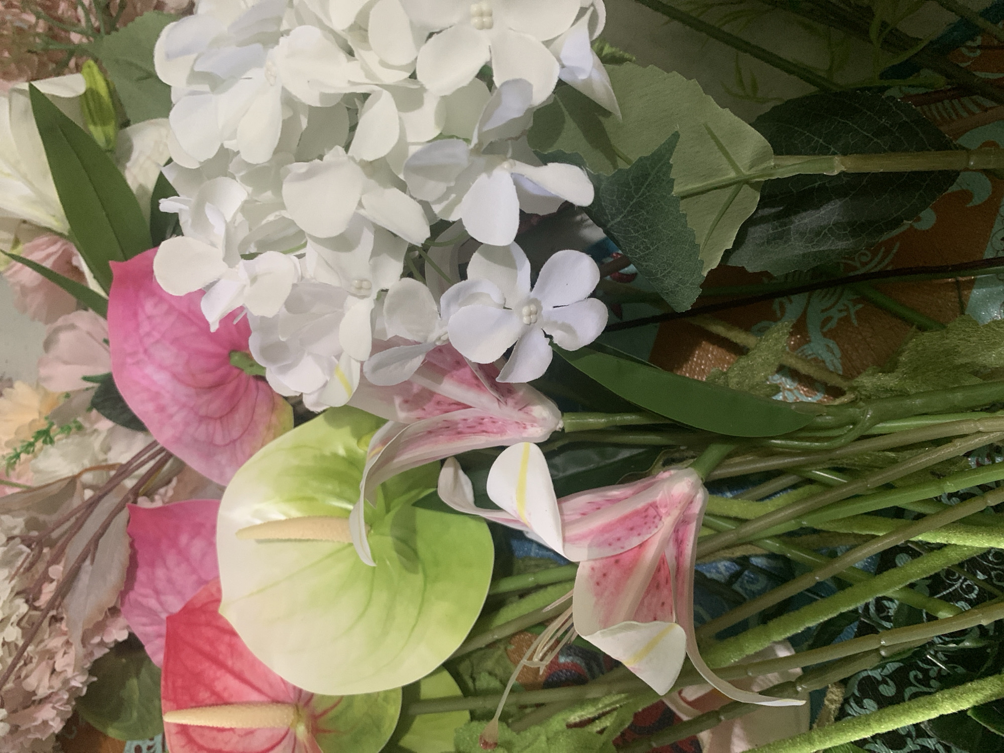 The image shows a close-up, overhead view of a floral arrangement. The arrangement features a variety of artificial flowers, including pink and red anthuriums, delicate pink and white lilies with speckled petals, and clusters of small white flowers that resemble hydrangeas. The flowers are supported by numerous green stems, some of which appear fuzzy. In the background, there are other floral elements in muted pink and cream tones, along with hints of greenery. The background surface is a mix of brown and turquoise, with a patterned texture. The lighting is diffused, creating soft shadows. There are no people or animals visible in the image, nor is there any indication of a specific location in Kuala Pembuang, Indonesia.