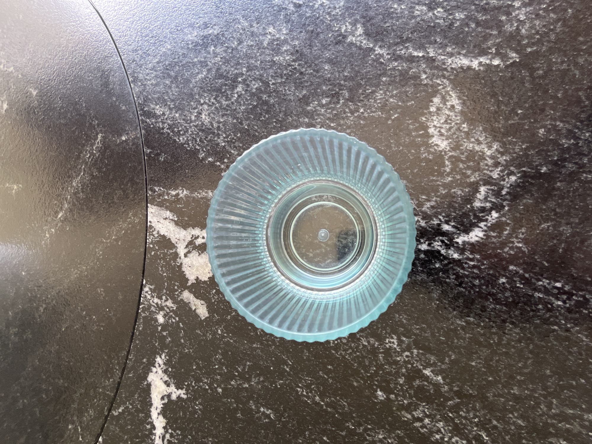 An overhead shot captures a translucent, light blue circular object resting on a dark, speckled surface. The object has a fluted or ribbed outer edge and a smooth, slightly raised inner base, suggesting it could be a coaster, a small dish, or the base of a drinking glass.
The surface beneath the object is dark grey or black with irregular white and light grey speckles and streaks, resembling a granite or quartz countertop. A prominent curved line runs from the top-left to the bottom-middle of the image, dividing the surface into two sections. The area to the left of this line appears slightly smoother and less speckled, or perhaps has a different texture, compared to the more visibly textured and patterned area to the right where the blue object sits. The lighting suggests an indoor setting. There is no activity taking place, no people, and no visible text.