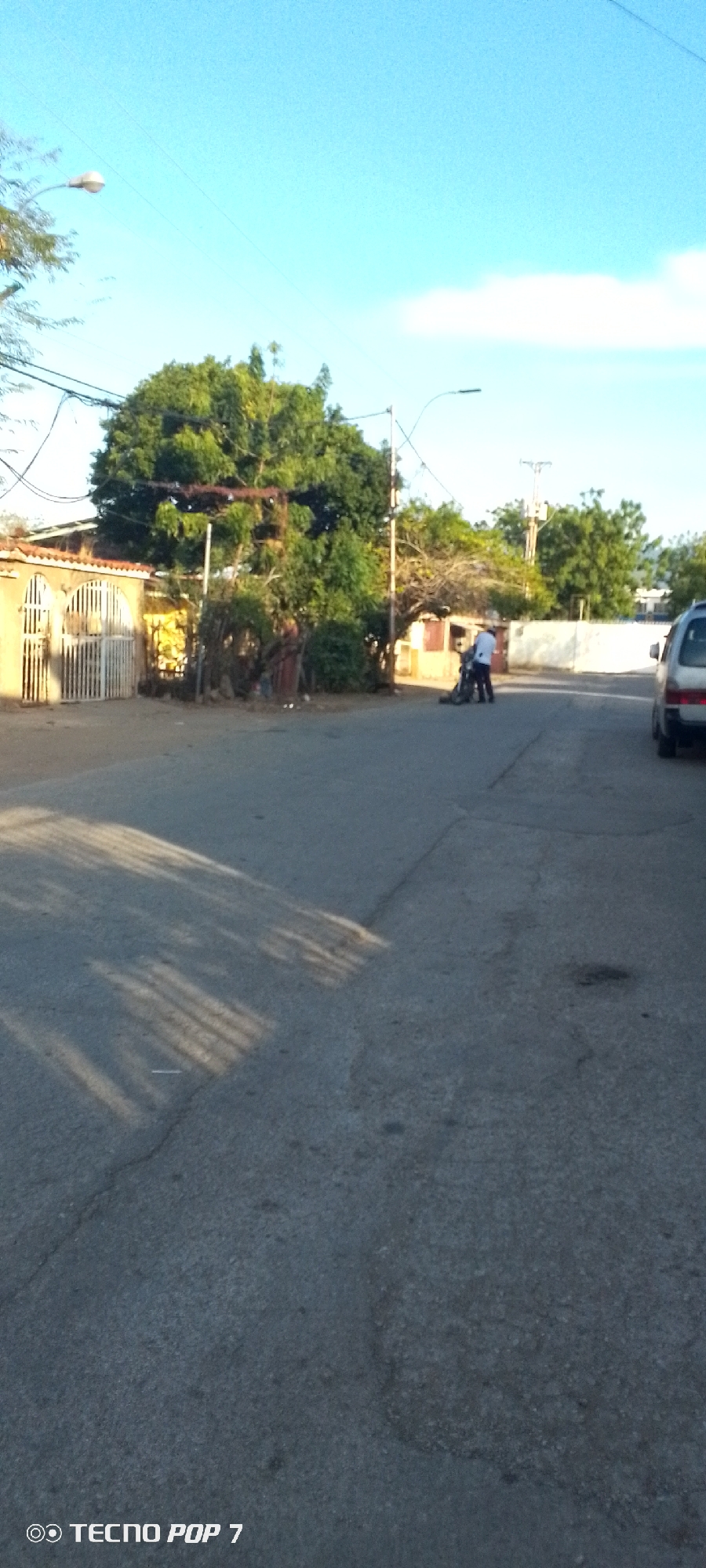 The scene depicts a residential street in Altagracia, Venezuela. The road, made of asphalt with cracks and shadows cast by trees, runs through the center. On the left side of the road, there are houses with fences and trees. On the right, a van is partially visible. In the distance, two individuals appear to be working on a motorbike. Overhead, a blue sky is visible. Streetlights are present. A "Tecno Pop 7" watermark appears at the bottom of the frame.