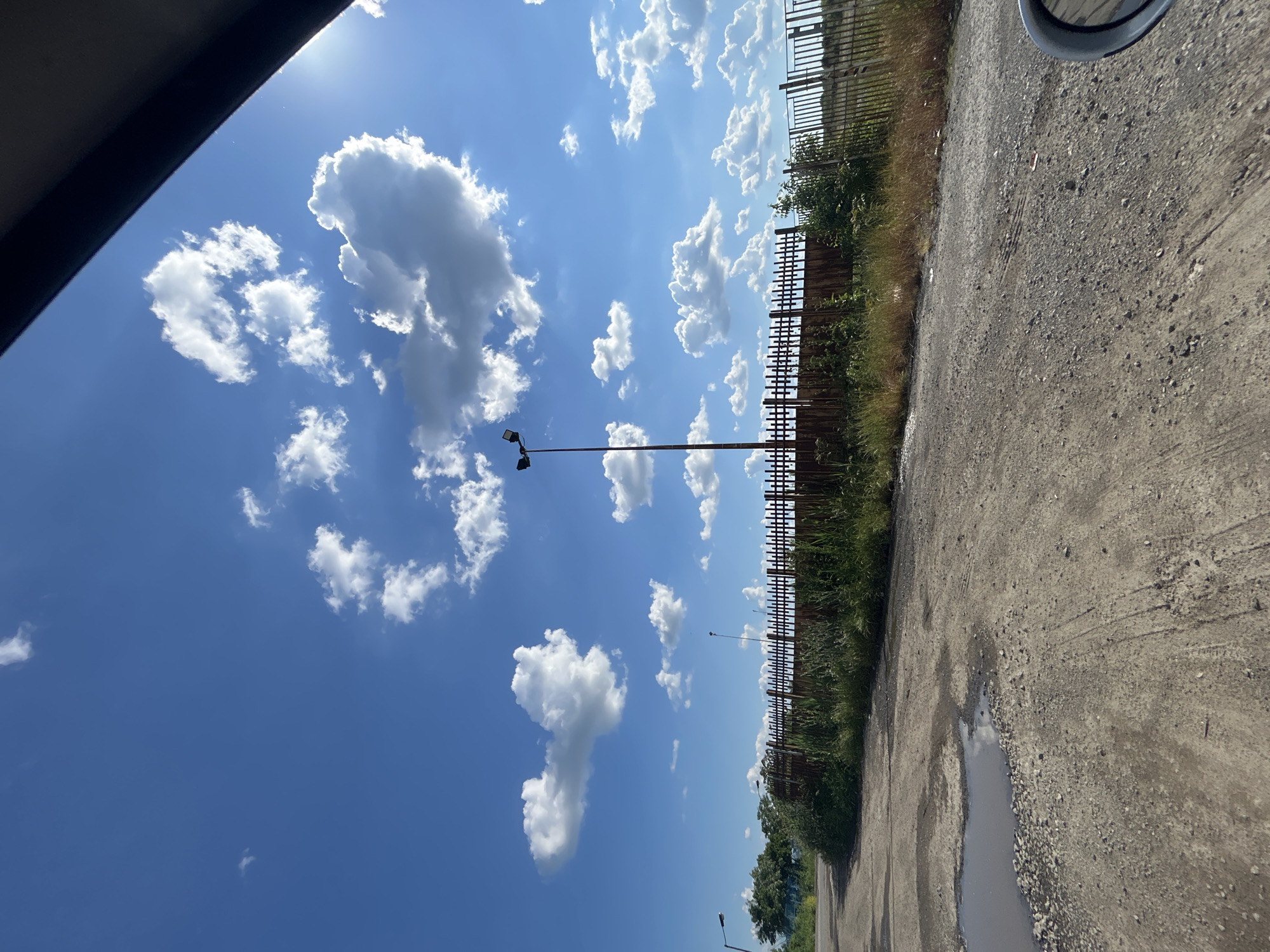 The image shows a view of a road and a blue sky with white clouds. A fence runs along the side of the road. A puddle is present on the road surface. Street lights are visible. Part of a vehicle, possibly a car, is visible at the edges of the frame.