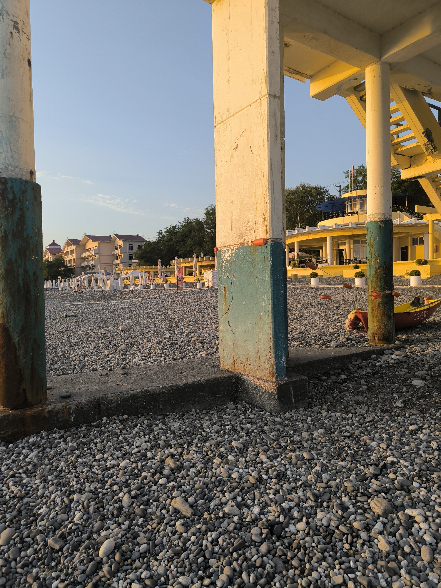 The image depicts a pebble beach in Sochi, Russia, on a clear day with soft sunlight casting long shadows. In the foreground, a concrete structure with thick pillars and weathered teal and rust paintwork dominates the scene, creating a framed view of the beach. The beach itself is covered in smooth, grey and white pebbles.
Beyond the foreground structure, rows of white beach umbrellas and loungers are set up, suggesting a public beach area. In the midground, a yellow building with an awning and white railings stands out, possibly a cafe or beachside establishment. A small red and yellow boat rests on the pebbles near the building, with a black cat perched on its side.
Further back, several multi-story buildings with red-tiled roofs are visible, set against a backdrop of lush green trees. The sky is a clear, pale blue, with a few wispy clouds. The overall impression is one of a serene, late afternoon at a seaside resort.