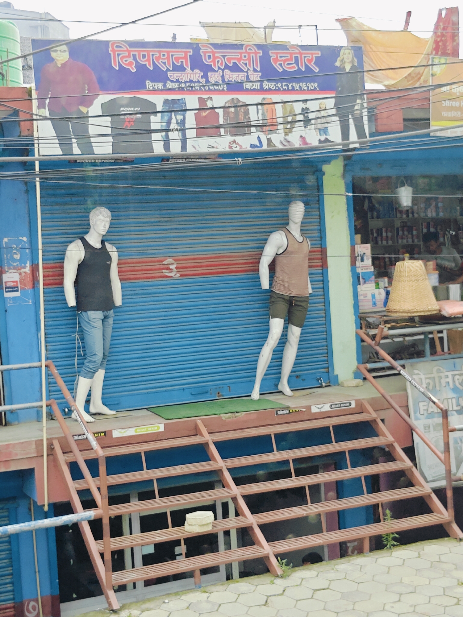 This image depicts the storefront of a clothing store called "Dipson Fancy Store" in Chandragiri, Nepal. The store features a blue rolled-up shutter and a blue exterior wall. Two mannequins are displayed outside the store on a raised platform with a metal staircase leading up to it. The mannequin on the left is wearing a dark tank top and light blue jeans. The mannequin on the right is wearing a striped tank top and dark shorts. A sign above the store displays the store's name in blue lettering. The surrounding environment includes electrical wires, other storefronts, and a paved sidewalk.