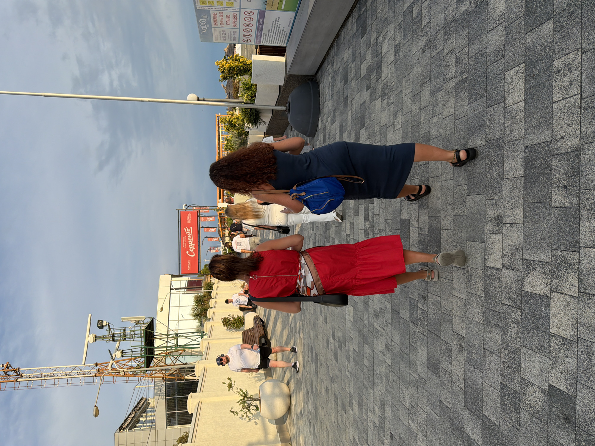 The image shows two women walking on a paved walkway. The woman in front is wearing a red dress and the woman behind her is wearing a dark blue dress. They are walking in the same direction. There are other people in the background, some of whom are walking and some are standing. In the background, there is a building with large windows, a crane, and several signs. One sign is in Russian and appears to be an informational sign about swimming restrictions, stating "ATTENTION! SWIMMING PROHIBITED 08:00-20:00". The scene appears to be an outdoor public space, possibly a promenade or a pedestrian zone. The lighting suggests it is daytime, with the sun casting shadows. The sky is partly cloudy. The overall mood seems casual and relaxed, consistent with a public outdoor setting. The text "Сорренто" (Sorrento) is visible on a red sign, possibly indicating a business or a place. The presence of a crane suggests ongoing construction or development in the area.