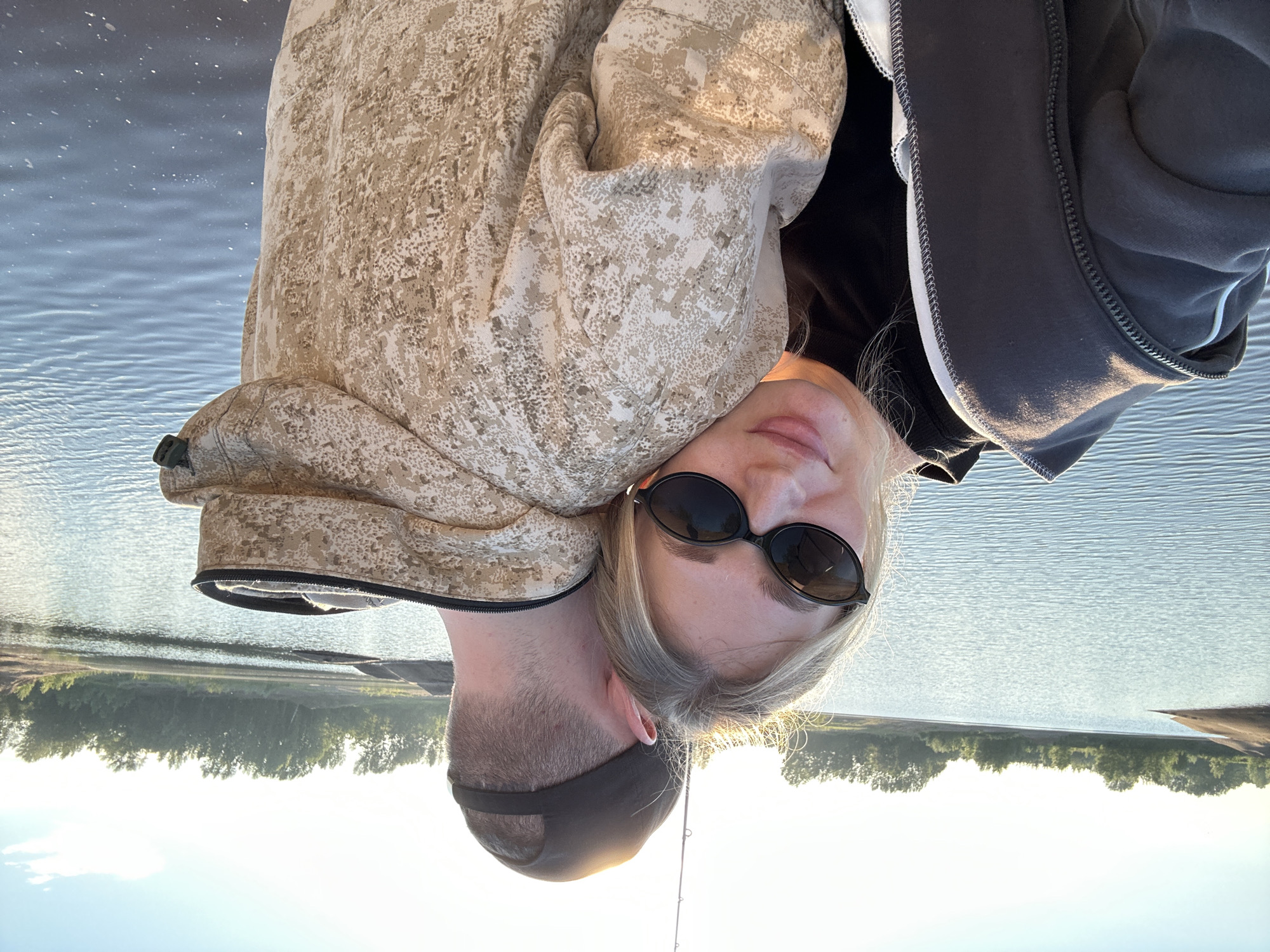 This image shows a couple embracing near a body of water, possibly the Kama River or a reservoir near Chaikovsky, Russia, given the surrounding trees and flat landscape. The woman, in the foreground, is leaning her head on the man's shoulder and looking towards the camera. She is wearing a dark top and a grey zip-up hoodie, with dark, oval-shaped sunglasses. Her blonde hair is visible around her face, catching the sunlight. The man, seen from the back, is wearing a camouflage hoodie and a dark baseball cap. A fishing rod is visible above his head, suggesting they might be fishing. The water is calm, reflecting the clear, bright sky, and the shoreline is lined with trees. The overall lighting indicates it is likely late afternoon or early evening, with a warm, golden hour glow.