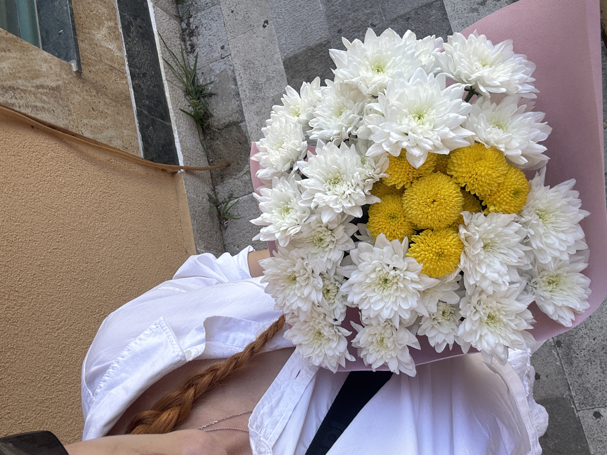 A medium shot, from slightly above, shows a person holding a bouquet of white and yellow chrysanthemums wrapped in pink paper. The person wears a white button-down shirt and has a red-braided hair visible on their shoulder. A thin silver necklace rests on their chest. A black strap is visible, possibly from a bag. The setting appears to be an outdoor urban environment, evidenced by the presence of concrete steps and walls. Based on the style of architecture and streetscape, the location is consistent with an area in the city of Budva, Montenegro.