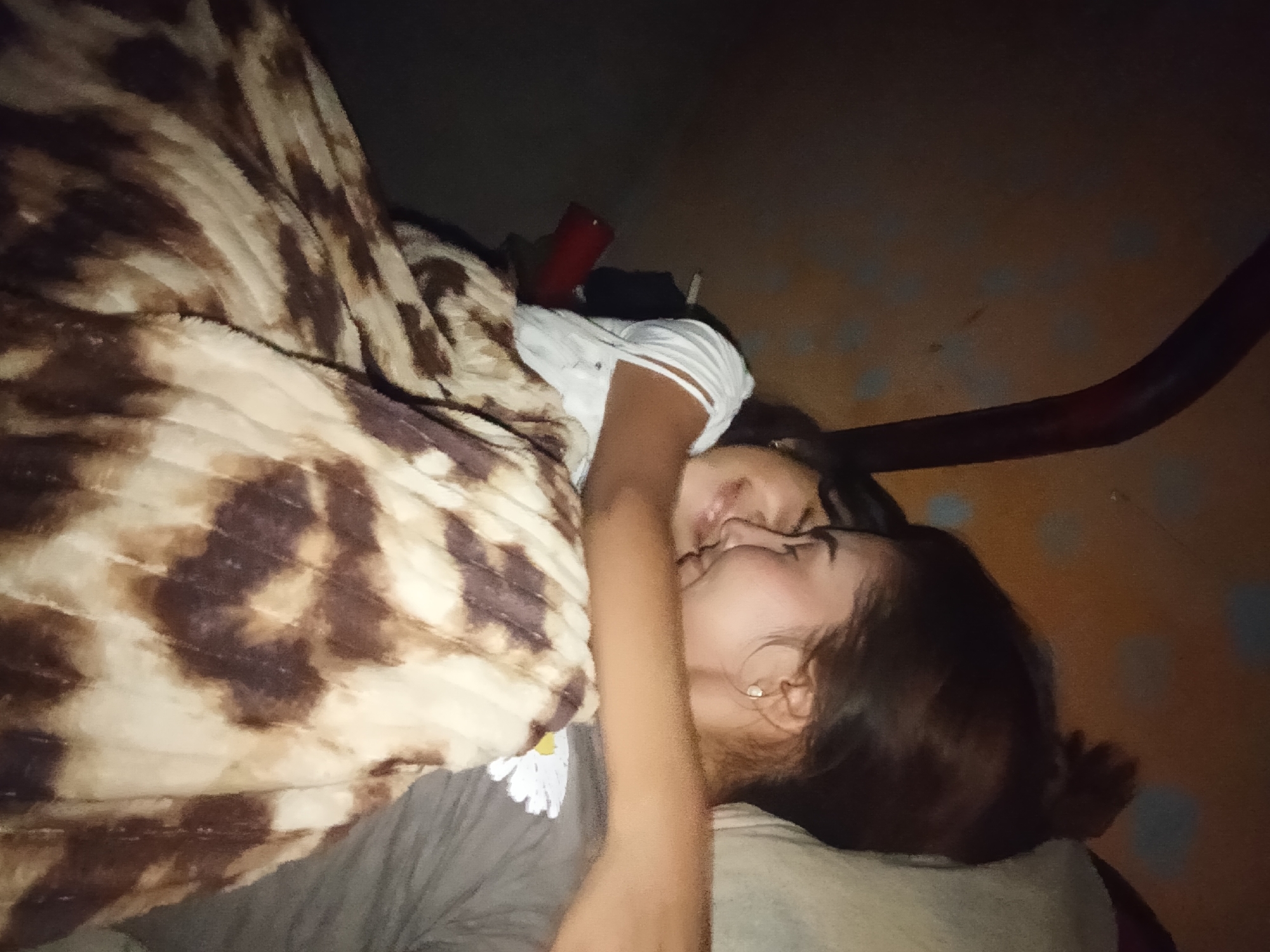 Two women are lying down, possibly on a bed or floor, covered by a patterned blanket. They appear to be kissing or nuzzling each other. One woman is wearing a gray shirt with a flower design, while the other is wearing a white shirt. The location is uncertain, though specified in the prompt as Juan Griego, Venezuela. There are some objects in the background including what seems to be a red cup and possibly a metal or wooden pipe. The lighting is dim.