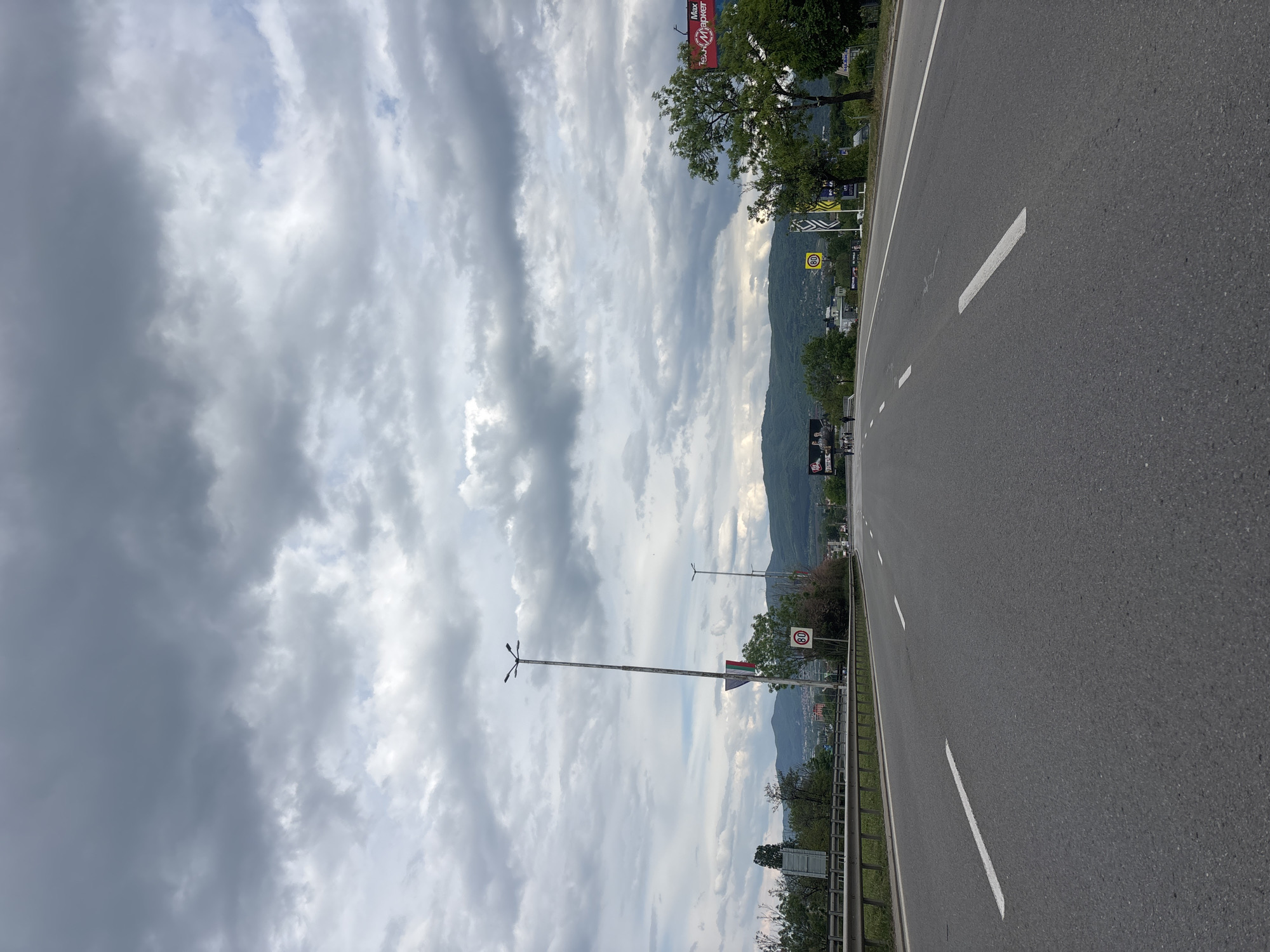 The image depicts a roadside view in Sofia, Bulgaria. The road is asphalt, with white lane markings. There's a metal guardrail separating the road from the scenery. In the distance, there are mountains visible. The sky is filled with various cloud formations. Road signs are visible near the road, including speed limits.