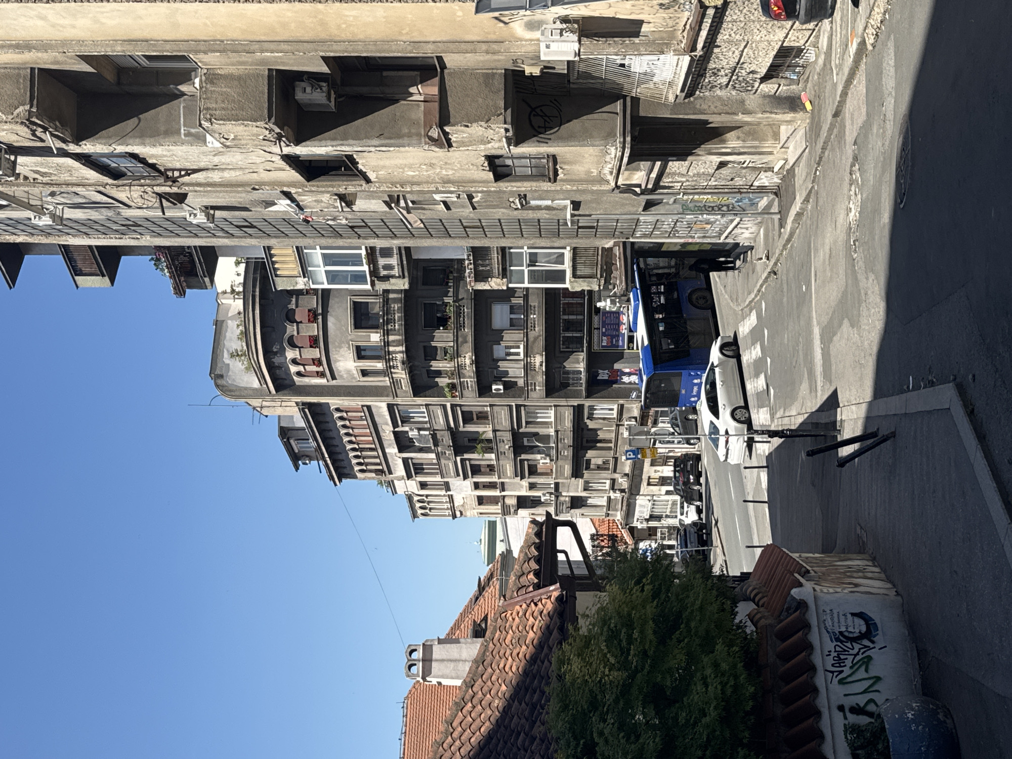 The image depicts a street scene in Belgrade, Serbia. The perspective is from a slightly elevated position, looking down a street lined with multi-story buildings. The architecture is predominantly from the mid-20th century, with some older structures featuring red-tiled roofs on the left.
The street is paved with asphalt and shows signs of wear and tear, with some visible patches and cracks. There are several vehicles present, including a blue public bus, a white sedan, and other cars parked or in motion. A pedestrian is visible on the sidewalk near the bus.
The buildings on the right side of the street are weathered, with peeling paint and exposed concrete. Many have balconies and windows, with some showing air conditioning units. The building on the far right is taller and appears to be in a state of disrepair.
On the left side of the street, a building with a red-tiled roof is partially visible, with a section of wall covered in graffiti. A green tree with dense foliage is also present in this area.
The sky is clear and blue, indicating a sunny day. Shadows cast by the buildings suggest the sun is high in the sky. The scene conveys a sense of an urban environment with a mixture of architectural styles and varying states of upkeep.