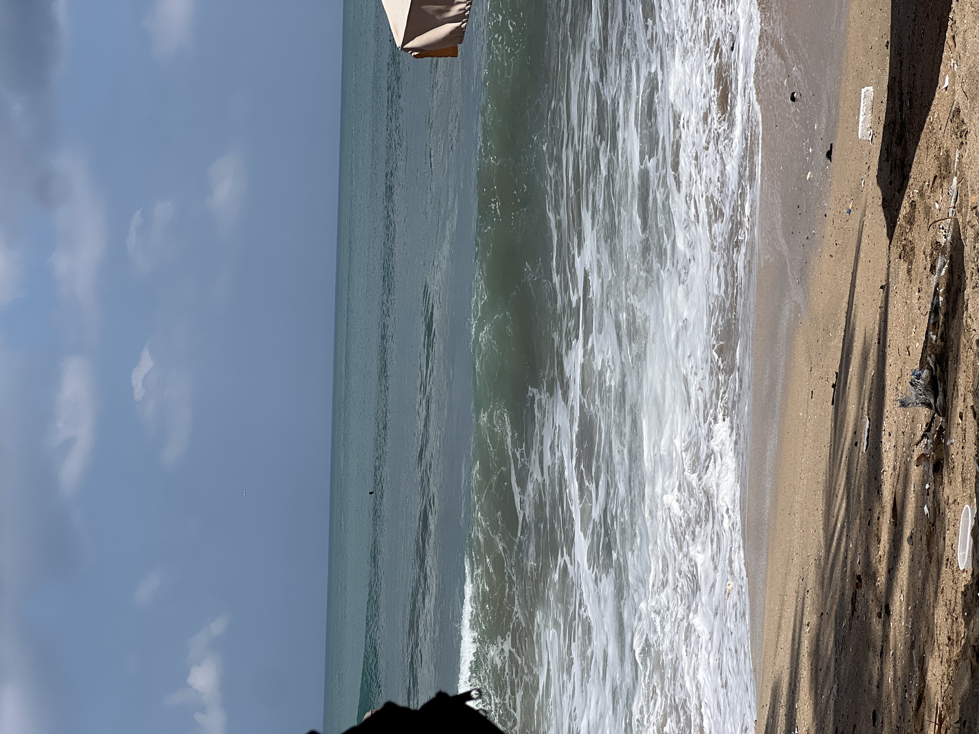 The image captures a scenic view of Kuta Beach in Indonesia. The foreground is a sandy beach with some debris scattered on it, including plastic waste and driftwood, casting shadows from unseen objects. In the middle ground, the ocean churns with white foam as waves break on the shore. A surfer is visible in the distance, riding a wave, and a distant airplane is in the sky. The background features the vast expanse of the ocean meeting the horizon under a partly cloudy blue sky. On the right side of the image, part of a beige beach umbrella is visible, suggesting a populated area. The overall impression is of a typical beach scene in Kuta, with its characteristic waves and sandy shores, but also showing signs of pollution.