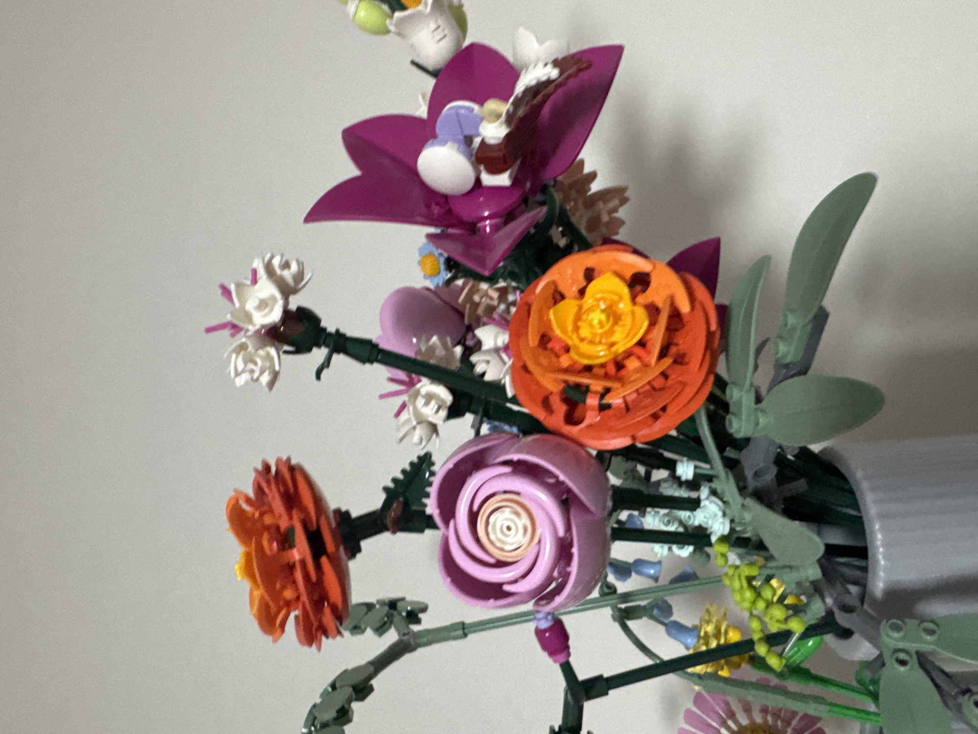 The image shows a close-up, eye-level, indoor shot of a bouquet of artificial flowers, likely made of LEGO bricks. The flowers are arranged in a grey vase and are in various colors including pink, orange, purple, white, and yellow. Some of the flowers have intricate petal designs. The background is a plain, light-colored wall. There are no people, animals, or any discernible landmarks that would indicate a specific location in Kyiv, Ukraine. The scene is static and focuses solely on the floral arrangement.