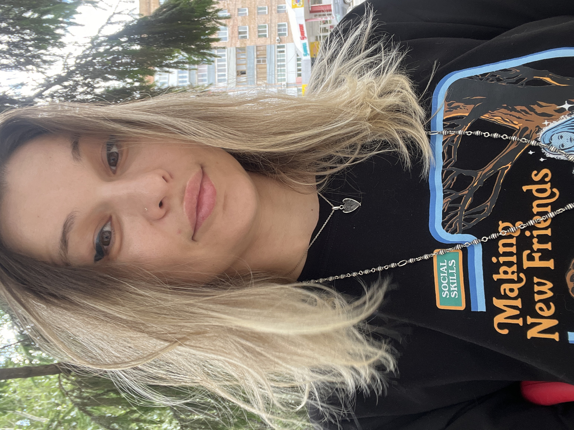 A medium shot of a young woman with blonde hair and a black sweatshirt is captured from the chest up. She is outdoors, with greenery and trees in the background, and a building with windows and balconies is visible further away, suggesting an urban setting. The woman has light skin, brown eyes, and light brown hair with blonde highlights, styled loosely around her face. She is wearing a black sweatshirt with a graphic print that includes text and imagery. A silver necklace with a heart-shaped pendant is visible around her neck. The overall lighting suggests daylight.