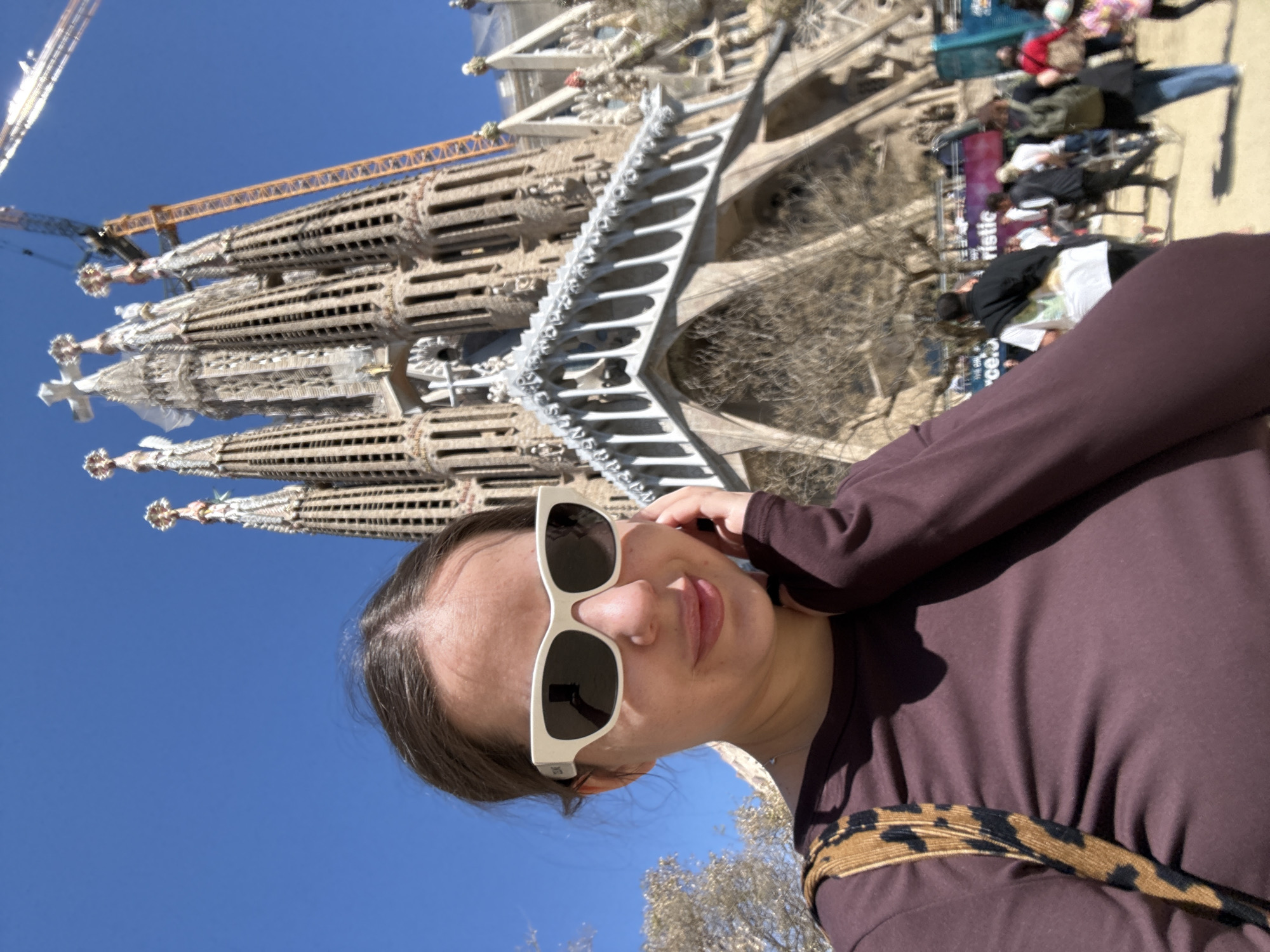 A woman wearing sunglasses and a brown long-sleeve shirt is facing the camera with a tilted head. Behind her is the Sagrada Familia in Barcelona, Spain, a famous basilica. There are people in the background observing the basilica. A construction crane is also visible. The sky is blue.