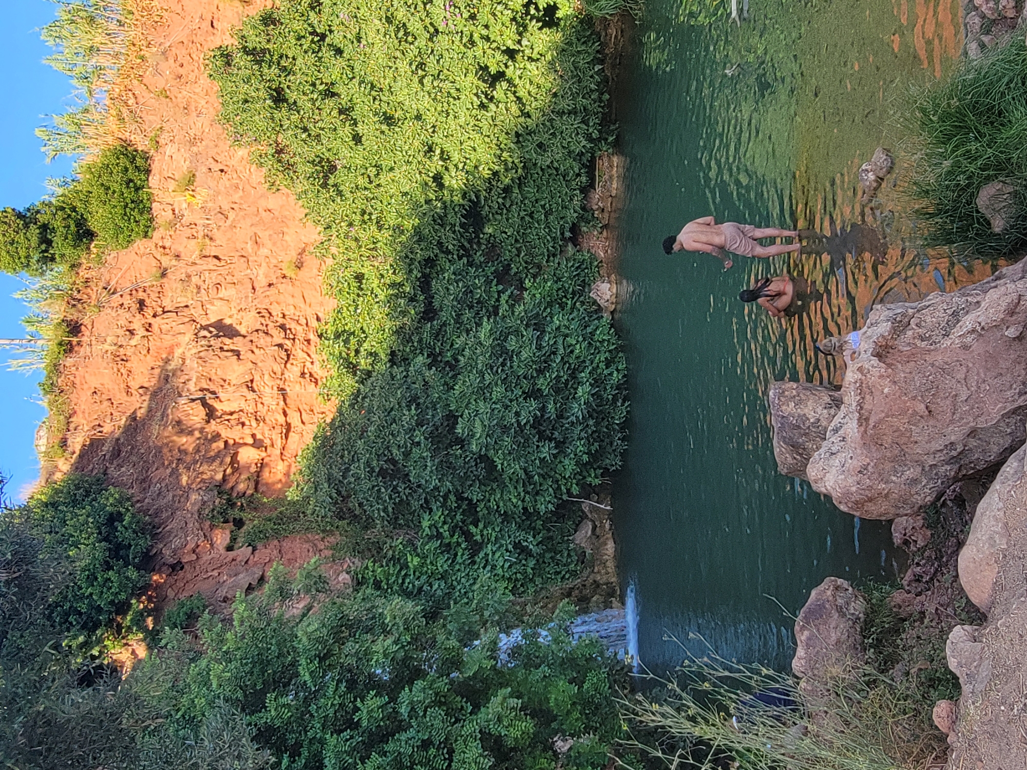 The image depicts a natural swimming area with clear green water, surrounded by lush vegetation and a rocky, reddish-brown cliff face. A small waterfall cascades into the water on the left side of the frame. In the water, two individuals are visible: a man standing and a woman seated. Another person is partially visible in the foreground. The scene suggests a recreational setting, possibly a natural pool or a designated swimming spot within a scenic landscape. The environment appears to be a warm climate, given the foliage and the presence of people swimming. The location is identified as being within the city of Loulé, Portugal.