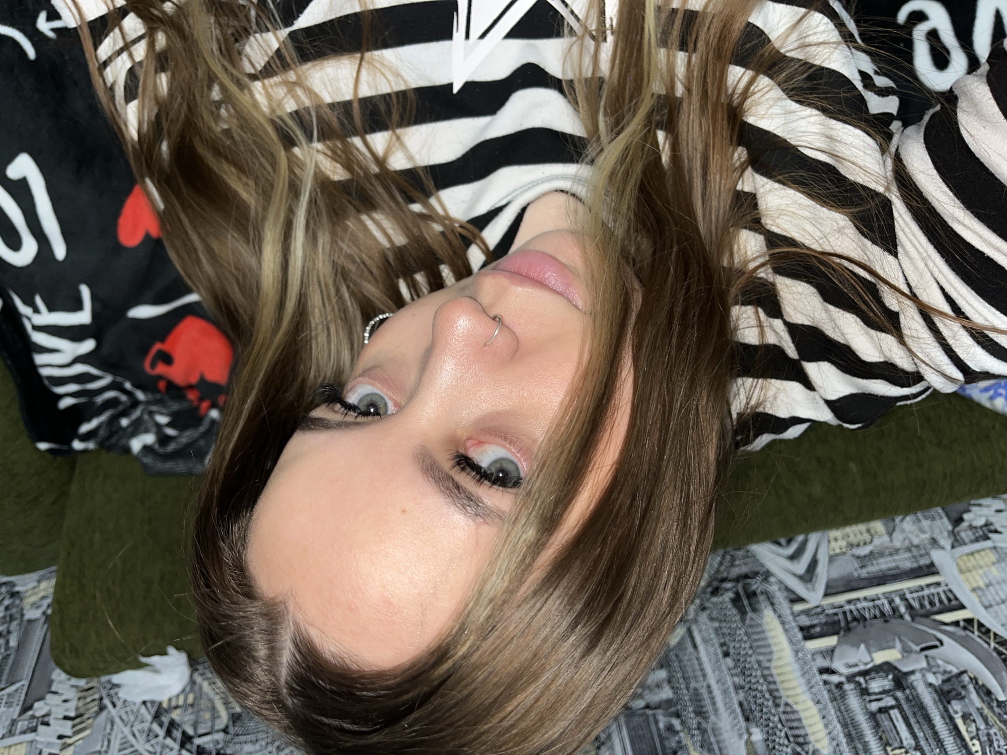 A close-up, slightly off-center, tilted shot shows a fair-skinned person with light brown hair, a nose ring, and light-colored eyes. They are wearing a black and white striped garment. Part of a black blanket with white writing and red heart/skull designs is visible on the left, and a patterned surface is seen on the bottom right. The location, Novopokrovskaya, Russia, is provided only as context but not discernible from the image itself.