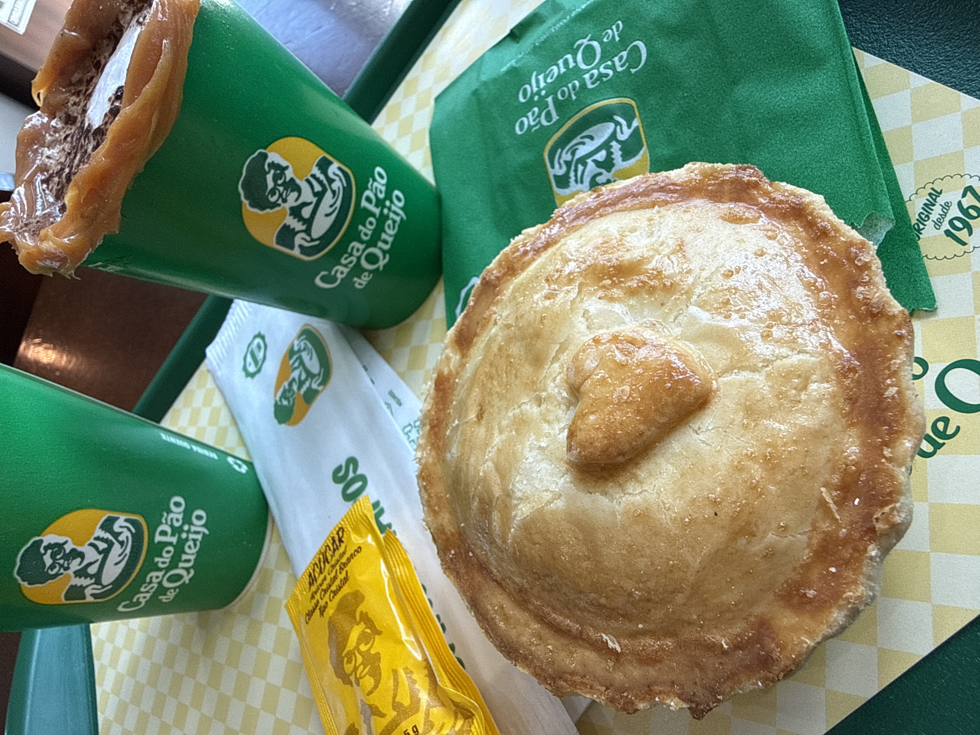 The image depicts a meal served on a dark green rectangular tray, presumably within a "Casa do Pão de Queijo" establishment in Guarulhos, Brazil. The tray is covered by a yellow and white checkerboard paper placemat featuring the "Casa do Pão de Queijo" logo (an illustration of an older man with glasses and a chef's hat, holding a plate of food) and the text "ORIGINAL desde 1967" in a scalloped design at the bottom right.
Central to the foreground is a round, baked pastry with a golden-brown, slightly flaky crust. A small, heart-shaped piece of dough is applied to the center of the pastry's top surface. The crust exhibits a glossy finish, likely from an egg wash or glaze, with subtle sugar crystal reflections visible along the edges.
Two dark green paper cups, bearing the same "Casa do Pão de Queijo" logo on a yellow circular background, are positioned towards the back of the tray. The cup on the left is partially visible, containing a dark beverage. The cup on the right is more prominent, holding a beverage lavishly topped with a thick, caramel-colored substance (consistent with *doce de leite* or dulce de leche) that drips over its rim. Beneath this caramel layer, a white, frothy substance, possibly whipped cream or milk foam, is visible, speckled with dark fragments, likely chocolate.
Adjacent to the left cup and the pastry is a small yellow sachet of sugar, clearly labeled "AÇÚCAR" (Portuguese for sugar), with additional text "Classic Cristal Branco" and "Fino Cristal," and a smaller version of the brand's character logo. A folded white paper napkin or sachet, also displaying the brand's logo, is positioned beneath the sugar packet. Another folded dark green napkin or piece of packaging with the brand's logo in white is visible beneath the right side of the pastry.
The background is softly blurred, showing warm, muted brown tones, suggesting an indoor commercial setting like a cafe or snack bar. No individuals are present in the frame. The items are arranged for immediate consumption.