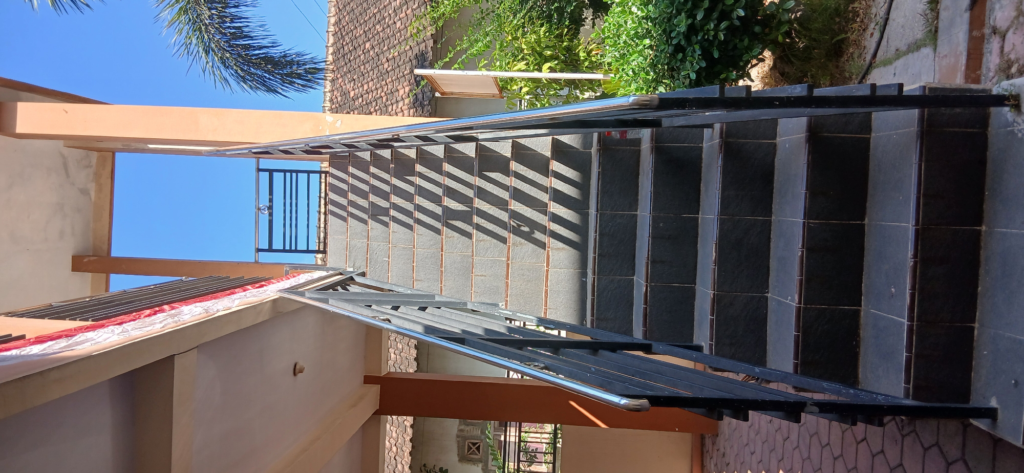 The image depicts an outdoor stairway in Pejaten, Indonesia. The stairway is constructed with dark grey tiled steps and features a black metal railing with a silver handrail. The steps are illuminated by sunlight, casting long, diagonal shadows across them. To the left of the stairs, a building with orange pillars and a light-colored ceiling is visible, adorned with a red and white banner. Behind the building, a tiled roof and greenery are present. To the right of the stairs, lush green bushes and a palm tree's frond are visible against a clear blue sky. The ground at the base of the stairs is paved with reddish-brown interlocking bricks. A single red plastic bottle rests on one of the steps.