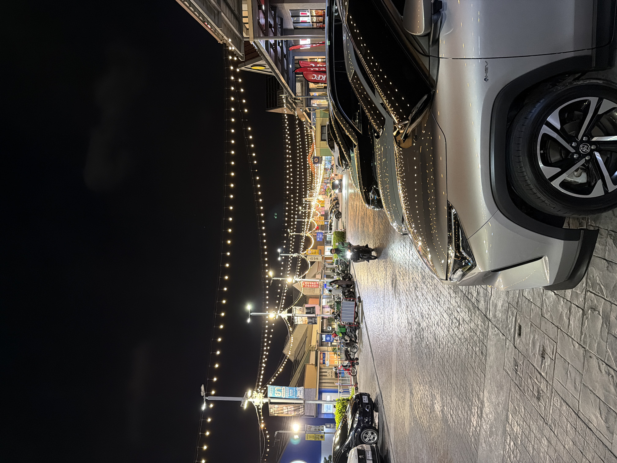 The photograph captures a nighttime scene in Pattaya City, Thailand. A row of vehicles is parked along a brick-paved street. The street is illuminated by overhead string lights, and the fronts of commercial establishments line the street behind the parking. Several motorbikes are parked nearby, and there are people present further down the street. The night sky is dark above.