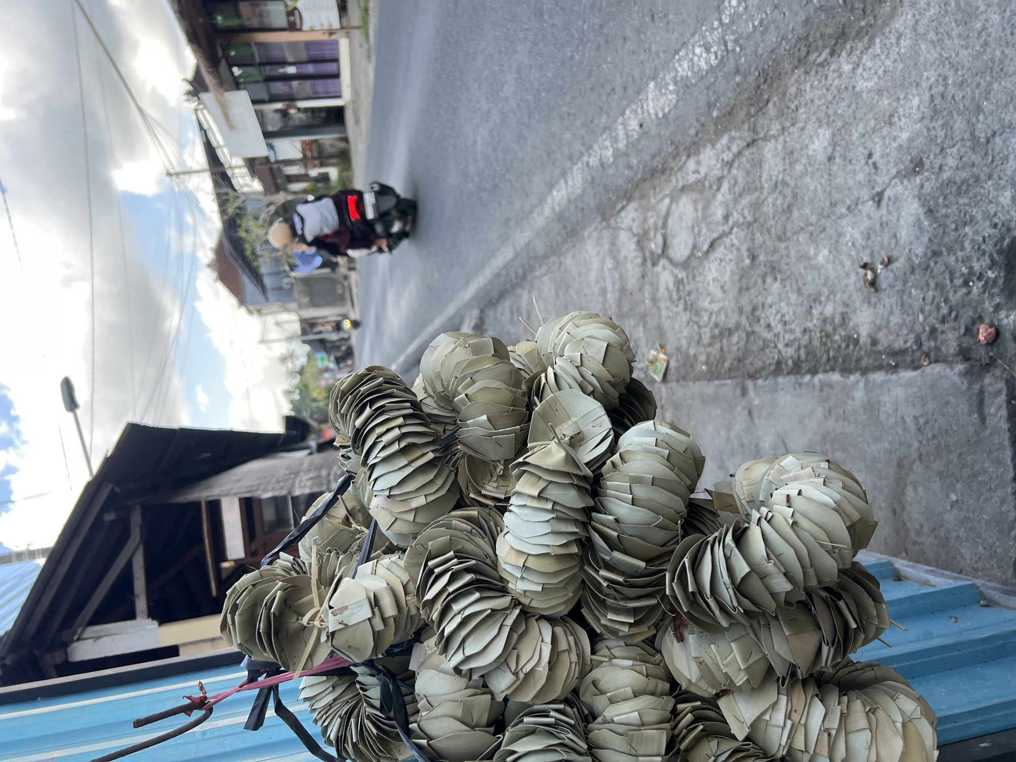 The photograph, taken in the Ubud District of Indonesia, presents a street scene with a focus on a large collection of woven palm leaf offerings arranged on a blue corrugated metal surface, likely a vehicle bed. These spherical or conical shapes are densely packed and dominate the foreground.
In the background, a street stretches into the distance, with buildings on either side. A person on a motorbike wearing a white helmet is riding away from the camera on the road. The sky is partly cloudy, suggesting daylight. The pavement shows signs of wear and repair.
