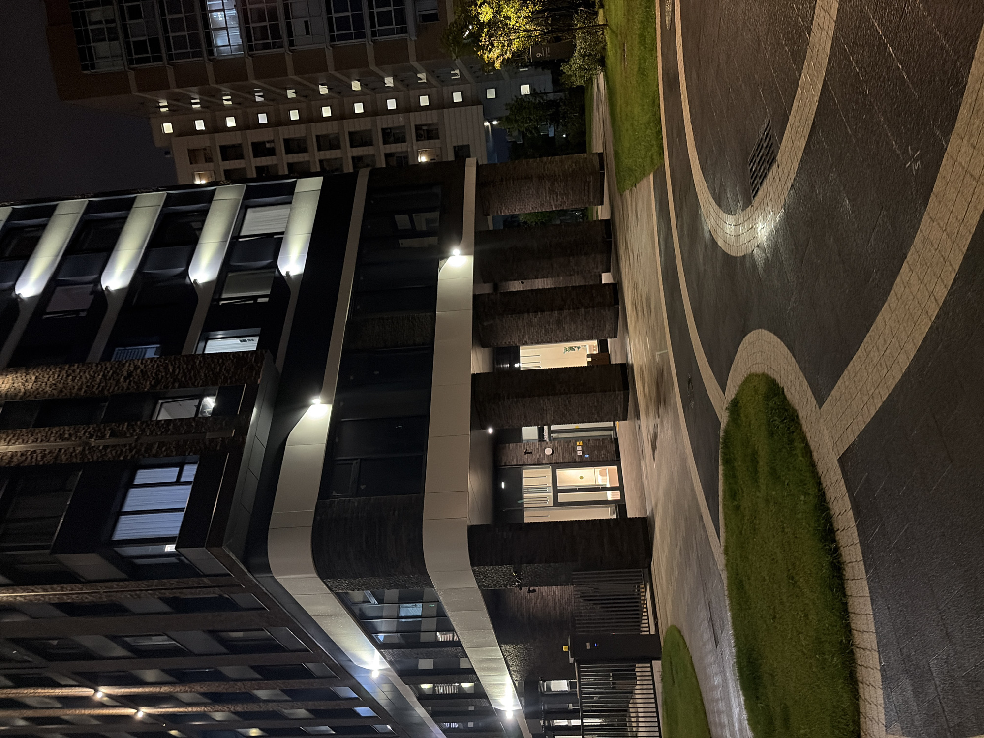 This image captures a modern residential building complex at night, located in Moscow, Russia. The scene is an outdoor shot focusing on the ground level entrance and surrounding landscaping of one of the buildings. The building itself is tall and contemporary, with dark brickwork and white trim. Exterior lights illuminate the facade, highlighting windows and architectural features.
In the foreground, a paved pathway with a distinctive curved design curves through the scene. The pathway is made of dark material with lighter colored inlaid patterns. To the left and right of the pathway are patches of green grass, which appear well-maintained. A dark metal fence with vertical bars is visible to the far left, suggesting a boundary to the property.
The entrance to the building is visible, featuring glass doors and an illuminated interior. Large, cylindrical pillars covered in dark brickwork support an overhang above the entrance. Several security cameras are mounted on the building's facade near the entrance.
In the background, another tall residential building with many illuminated windows is visible, indicating that the complex is occupied. A single tree with illuminated yellow foliage stands to the right of the building, adding a touch of color to the dark night scene. The overall atmosphere is that of a quiet, modern urban environment at night.