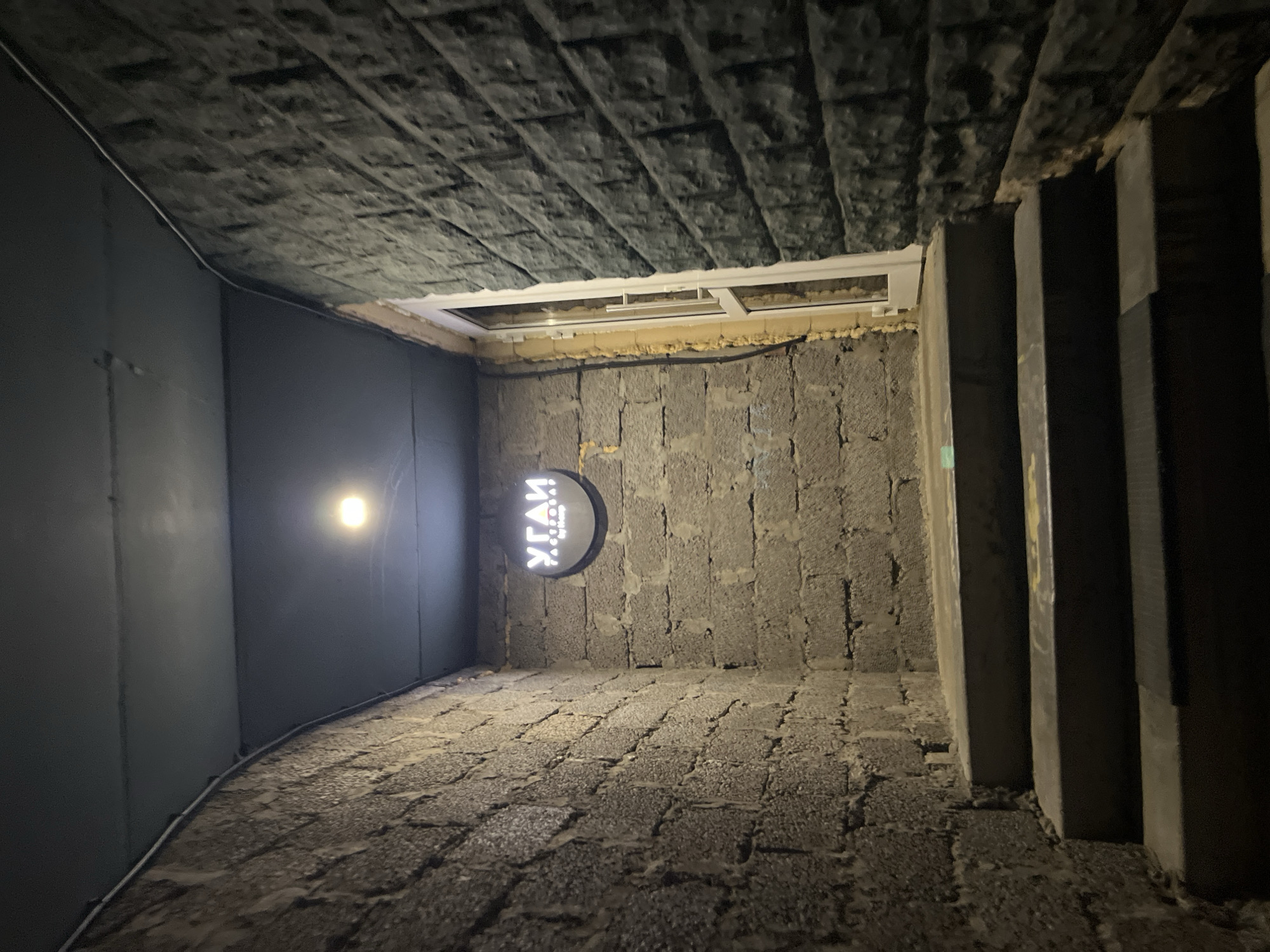 The image depicts a narrow, enclosed passage or entryway in Chaikovsky, Russia, viewed from a low perspective looking upwards. The scene appears to be set at night or in an interior space, lit by artificial light.
The left wall is covered with dark, smooth panels, along which a thin white cable or pipe runs near the ceiling. A single, prominent circular light fixture with a warm glow illuminates this wall and the textured floor below. The right wall is constructed from rough-hewn, dark grey stone or concrete blocks. Mounted on this wall is a circular, backlit sign featuring white Cyrillic text on a dark background. The discernible text reads "УГАИ" at the top, and below it, "УЛИЦА КУЛЬТУРЫ," which translates to "Street of Culture." To the far right, a dark, vertical structure resembling a staircase or a series of steps is partially visible.
The ceiling is covered with dark, textured panels, possibly insulation or acoustic material. Near the back, an open rectangular section with white framing, resembling a window or skylight, provides a glimpse of what might be an exterior or another level. The floor of the passage is paved with dark, irregularly shaped cobblestones or rough stone blocks.
The overall impression is one of a functional, possibly industrial or utilitarian, space with a rustic aesthetic due to the stone and cobblestone elements. The artificial lighting and enclosed nature create a somewhat intimate or mysterious ambiance. There are no people or specific activities depicted in the static image.