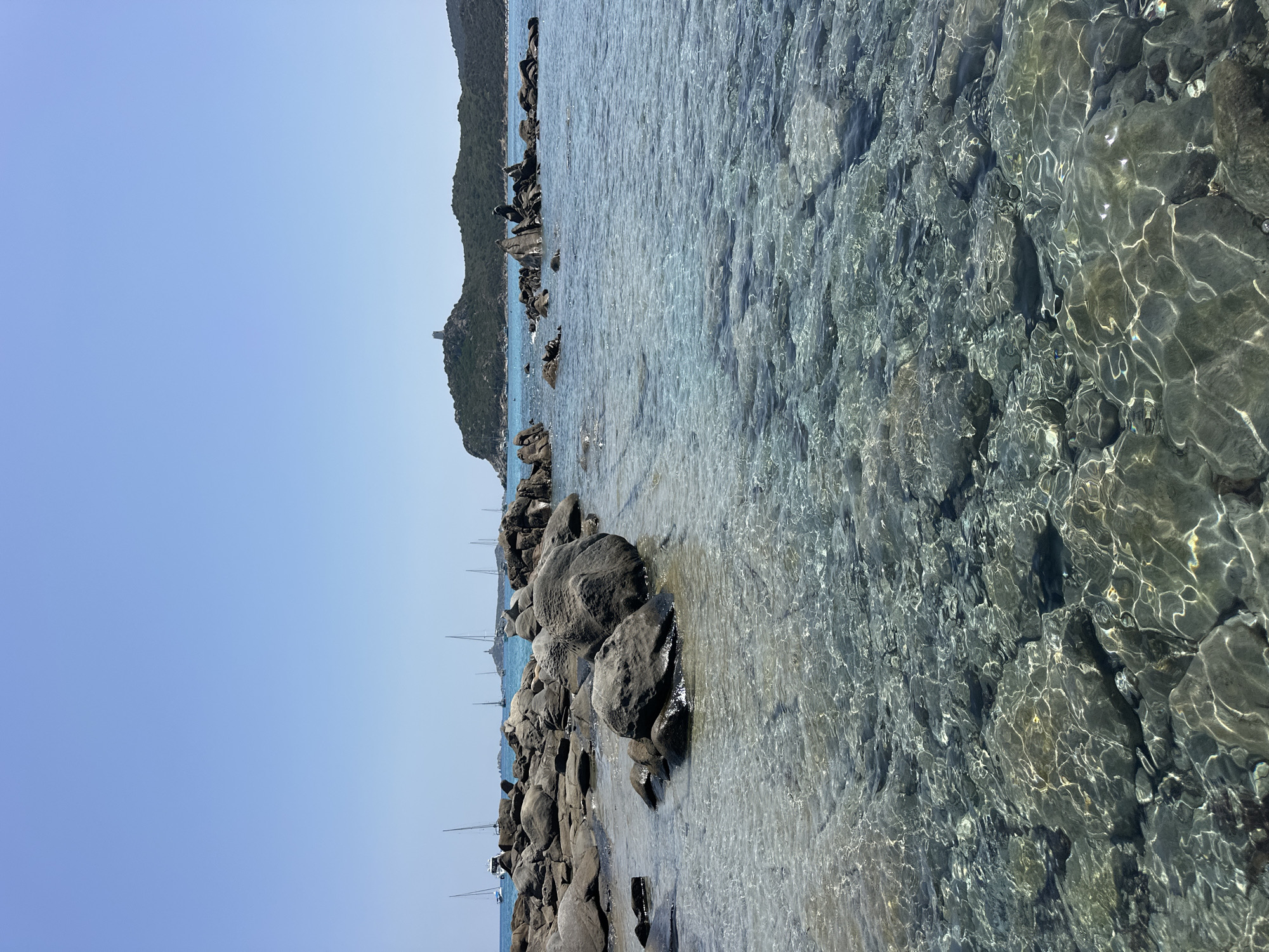 A scenic view of a clear turquoise sea with a rocky shoreline under a bright blue sky. In the foreground, large, dark gray rocks are partially submerged in the shallow water, with sunlight creating shimmering patterns on the seabed. Further out, several sailboats and a larger white boat are anchored or sailing on the calm water. In the distance, a hilly, tree-covered island is visible, topped with a small, stone tower. The overall impression is a tranquil coastal landscape, likely in a Mediterranean setting, possibly near Crabonaxa or Villasimius, Italy, known for its beautiful beaches and clear waters. There are no people visible in the image.
