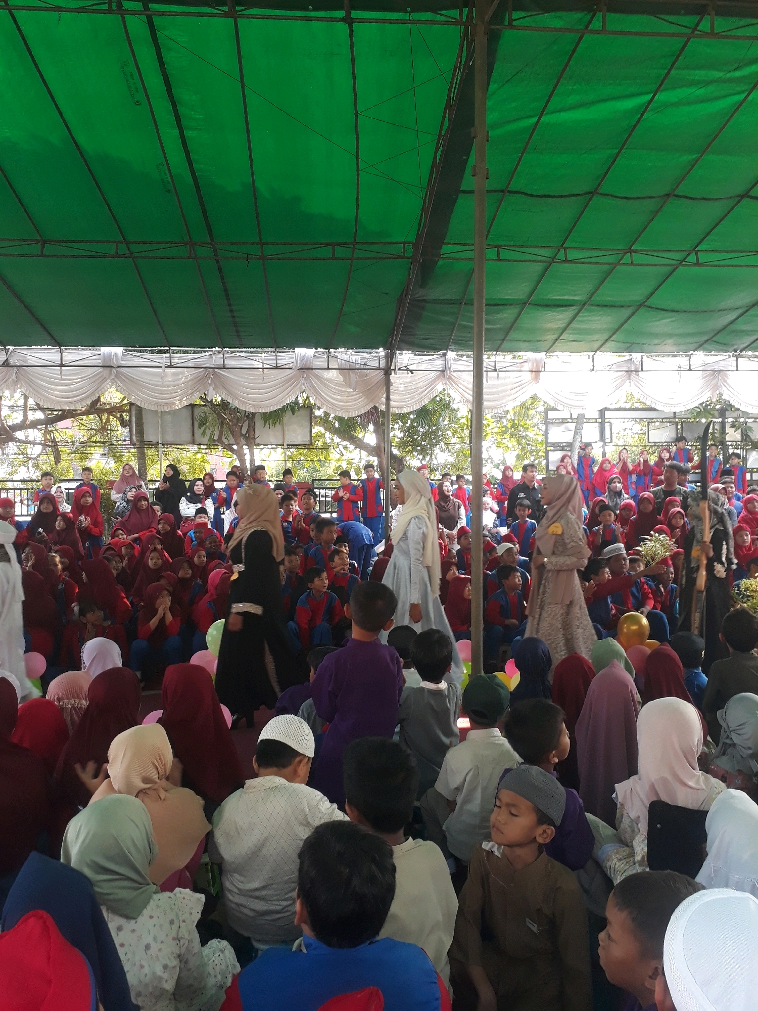 The image depicts a gathering of people, primarily children, seated on the ground outdoors under a large green tarp supported by metal poles. The setting appears to be a school or community event, possibly a religious ceremony given the attire of some individuals, including headscarves and caps. The children are dressed in school uniforms, predominantly red and blue. Several adults are also present, some standing and observing. In the background, there is a white draped curtain or backdrop, and beyond that, trees and buildings suggest an urban or suburban environment. There are also balloons visible in the foreground, adding a festive element. The lighting suggests it is daytime. The image was captured at SD Negeri 2 Pangkalpinang, located in Pangkalpinang City, Indonesia.