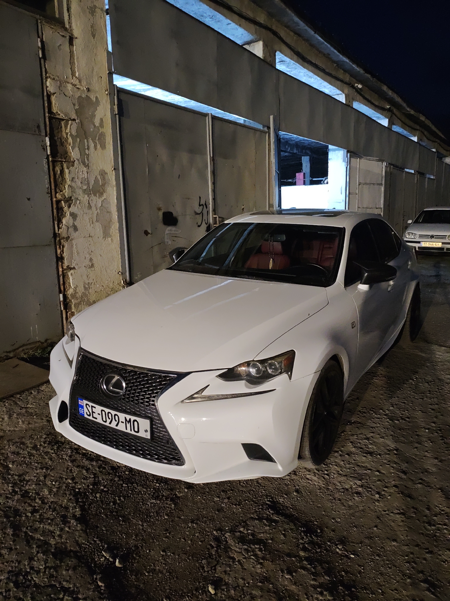 The image depicts a white Lexus sedan parked on a dirt lot in front of a large, industrial-style building with metal doors. The building appears to be a warehouse or garage. The car's license plate is visible and reads "SE-099-MO". Another car, a white Volkswagen Golf, is partially visible in the background. The scene appears to be at night, with artificial lighting illuminating the area. The ground is uneven and covered in loose gravel and dirt. The building's facade is weathered and shows signs of age. The overall impression is one of an urban or industrial setting. There are no people visible in the image. The location is stated to be Krtsanisi, Georgia.