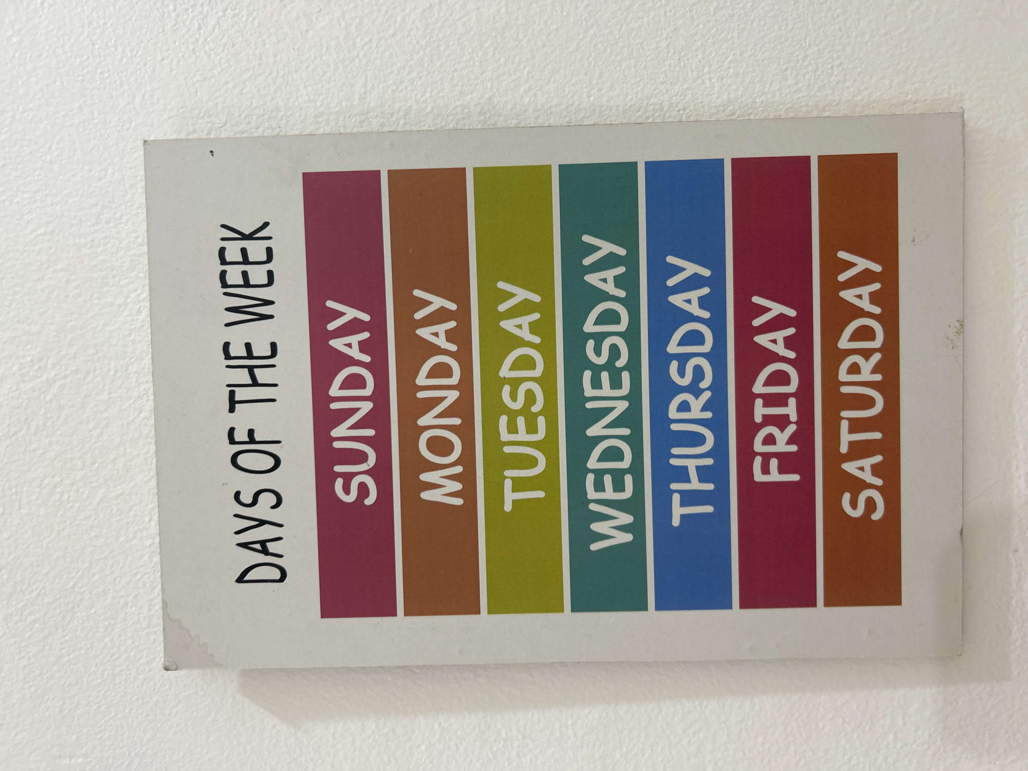 A sign with the days of the week is mounted on a textured white wall. The sign has a white border and a white background. The title "DAYS OF THE WEEK" is written in black, uppercase letters at the top of the sign. Below the title, the days of the week are listed in order, each on a colored background with white, uppercase letters. The colors are pink for Sunday, orange for Monday, green for Tuesday, teal for Wednesday, blue for Thursday, dark pink for Friday, and brown for Saturday. There are no people or actions depicted in the image. The environment is an interior wall. The location of Kuta, Indonesia is not visible in the image.