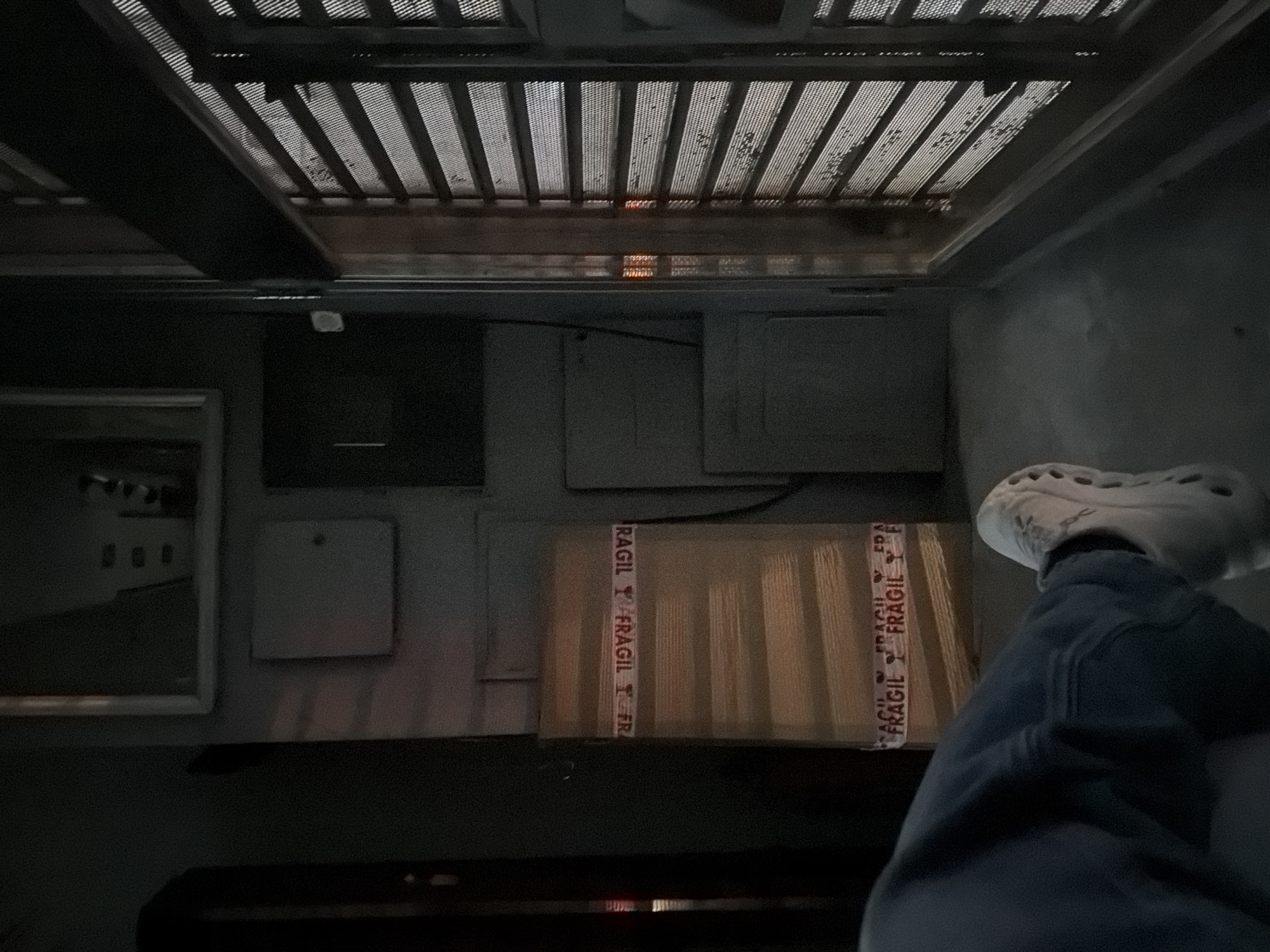 The image depicts an interior scene with a low-angle view upwards towards a ceiling with a grid-like structure. Below the ceiling are various rectangular panels or fixtures on the wall. A cardboard box with "FRAGIL" tape is visible, partially obscured by a person's leg wearing blue jeans and a white Croc-style shoe. The left side of the image includes a framed object, possibly a painting or mirror. The scene is dimly lit. There are no explicit indicators of the location being in Buenos Aires, Argentina.