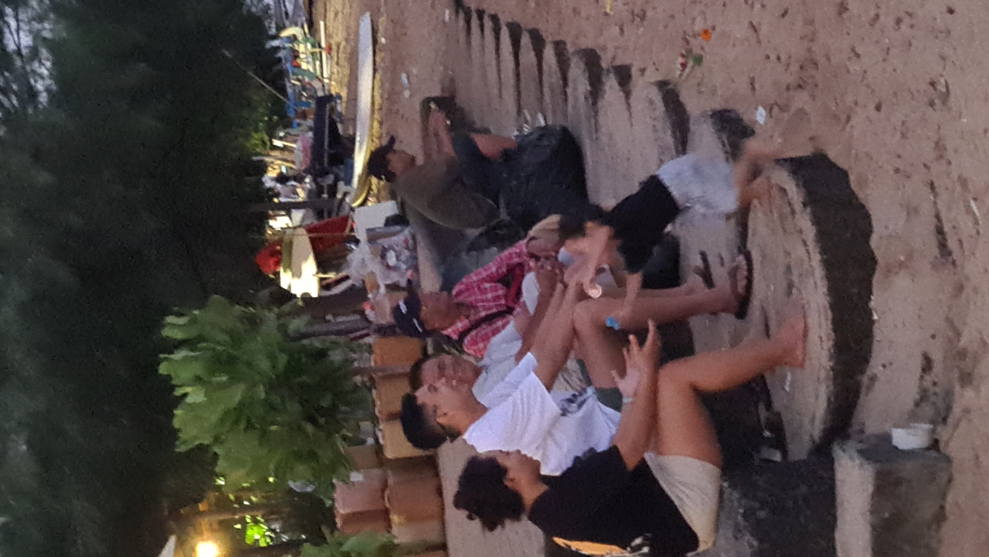 A group of people are sitting and interacting on a sandy area with concrete structures at the beach in Denpasar, Indonesia. In the foreground, a person in a black shirt with an orange emblem is seated on a concrete structure. To their right, two men are seated on similar structures, one in a white t-shirt and the other in a red plaid shirt and a dark cap. Further to the right, another man is seated, wearing a dark green shirt and a cap, and appears to be looking down at something in his hands. Between the seated individuals, a person is partially visible, possibly a child, with their legs extended and appearing to be reaching out. The background shows trees, foliage, some debris, and what appears to be beach equipment, including surfboards. The lighting suggests it is late afternoon or early evening.