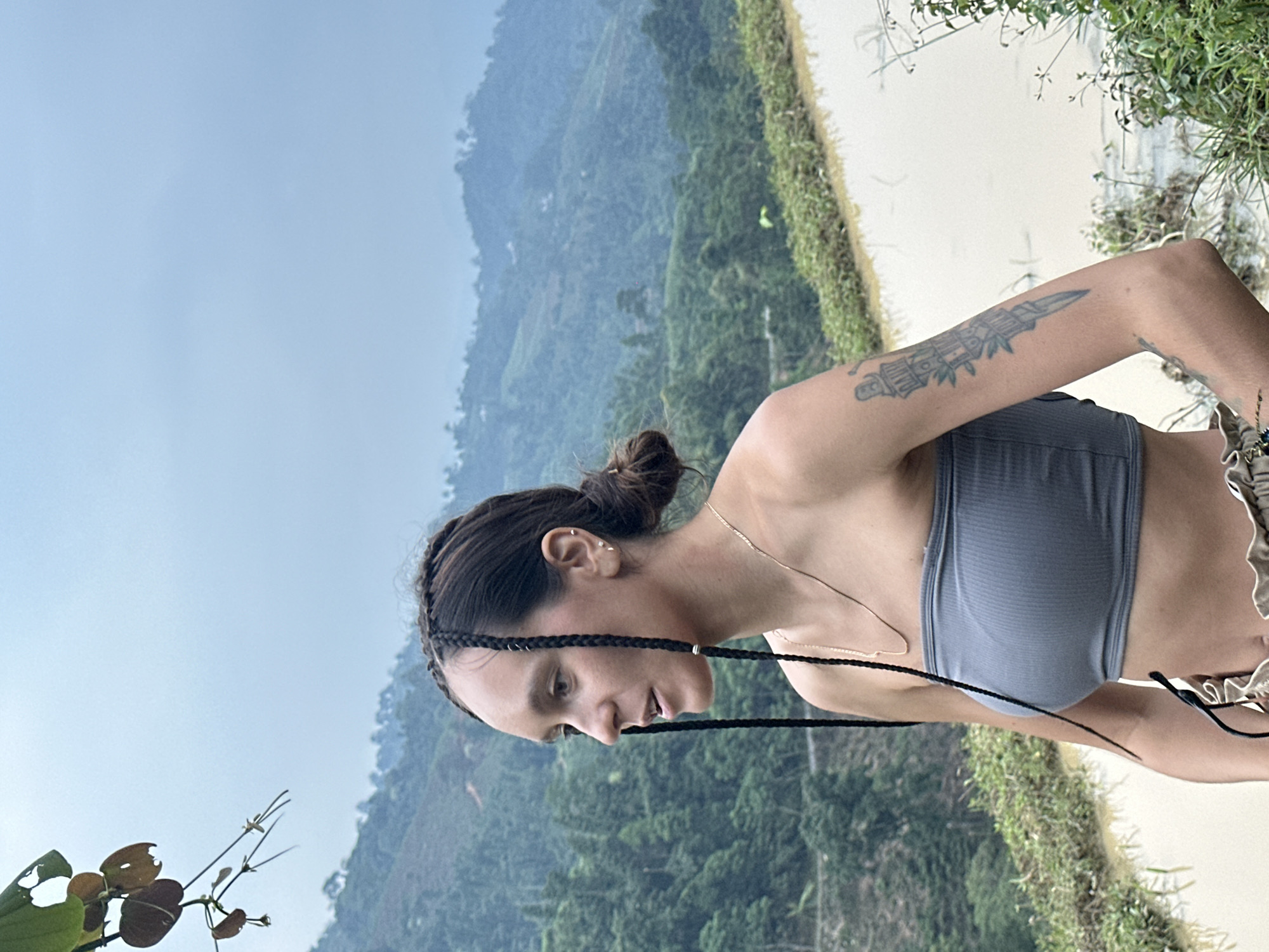 A young woman is depicted outdoors, looking downwards and to her right, against a scenic backdrop of mountains and water. She has dark hair, partially styled in small braids that frame her face and hang down, with the rest pulled back into a bun. She wears a grey ribbed crop top and a thin gold chain necklace. Several small stud piercings are visible on her ear. A notable tattoo, appearing to be an ornate dagger or sword design with intricate surrounding elements, is visible on her upper right arm.
The background features a vast expanse of lush, green, tree-covered mountains rising under a light blue, hazy sky. In the mid-ground and lower right, a calm, light-colored body of water, possibly a flooded rice paddy or a lake, reflects the sky. Green vegetation and grass line the edge of this water body. In the lower left foreground, some blurred leaves and branches are visible. The scene suggests a tranquil, natural environment during the daytime, likely in Nậm Dịch Commune, Vietnam. No discernible text is visible beyond the stylistic elements of the tattoo.