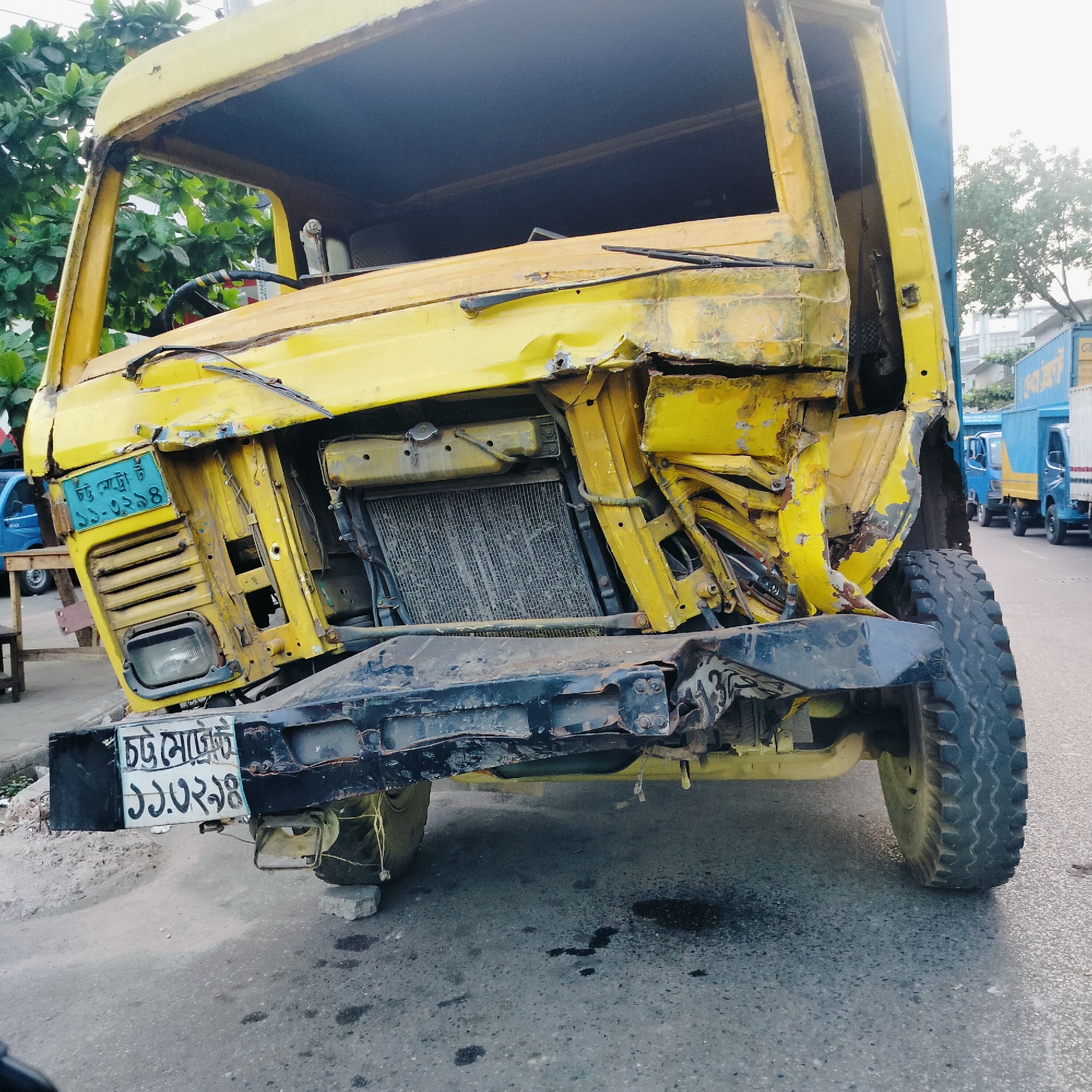 The image shows a badly damaged yellow truck in Unknown, Unknown. The front of the truck is severely crushed, with exposed radiator and twisted metal. The windshield is missing, and the license plate is partially obscured. The truck appears to be resting on concrete blocks. There is a puddle of liquid underneath the front. In the background, there are other trucks parked along the street. The environment appears to be an urban setting.