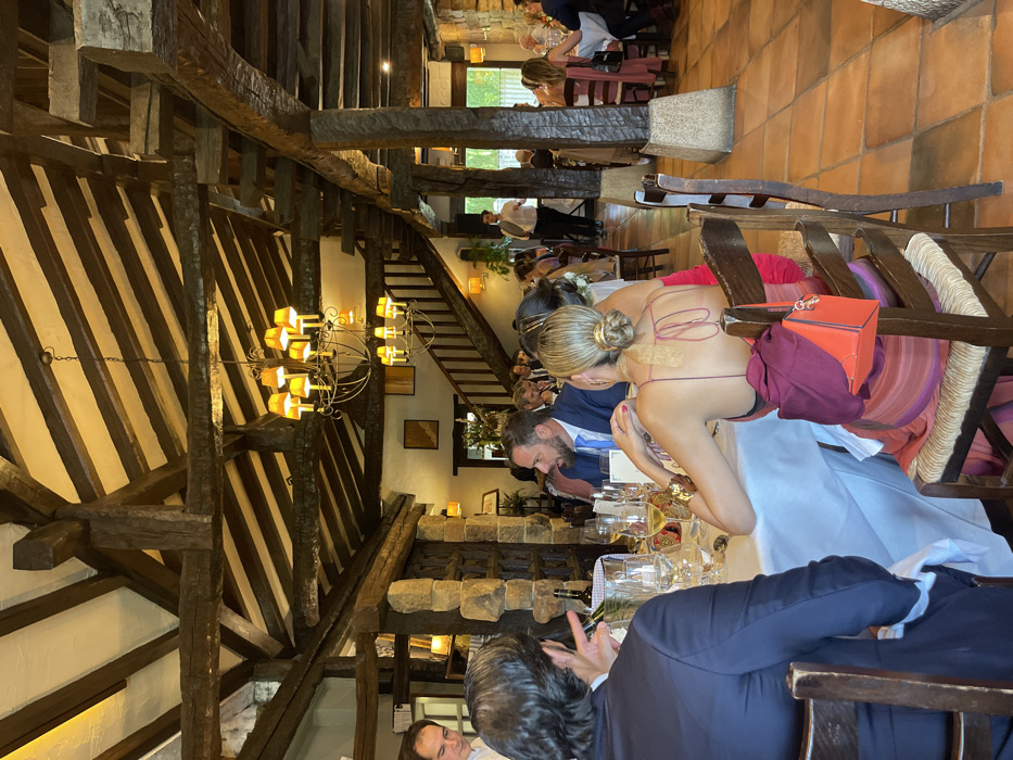The image captures an interior view of a large dining hall within a venue located in Amorebieta-Etxano, Spain. The architecture is characterized by a prominent ceiling structure of heavy, dark-stained wooden beams and support columns, some appearing clad with rough-hewn stone. Light-colored panels are visible between the ceiling beams. The flooring consists of terracotta tiles laid in a consistent pattern. Illumination is provided by multiple ornate metal chandeliers with yellow-shaded lamps, and additional wall sconces. A wooden staircase with railings ascends in the background, and an open entryway or large window is visible beyond.
The scene depicts a formal gathering, with numerous individuals seated at round and rectangular tables covered with white tablecloths. Most attendees are dressed in formal or semi-formal attire. In the immediate foreground, the back of a male wearing a dark blue suit jacket is visible on the left, with his right hand gestured as if speaking. To his right, the back of a female with blonde hair styled in a bun is shown; she is wearing a low-backed, maroon-colored dress with horizontal stripes towards the bottom. A reddish-orange purse and an additional piece of dark fabric are draped over the back of her wooden chair. At the same table, a bearded male in a blue suit and light blue tie is seated, looking towards the right.
Table settings include wine glasses, water glasses, plates, and cutlery. A silver wine cooler bucket containing a bottle is present on the foreground table, alongside glasses holding white wine. Staff members are observable; one is partially visible on the far left, and another in the background center-right, dressed in a white shirt and dark trousers, is moving between tables, appearing to serve guests. The overall activity involves guests seated, conversing, and partaking in a meal.