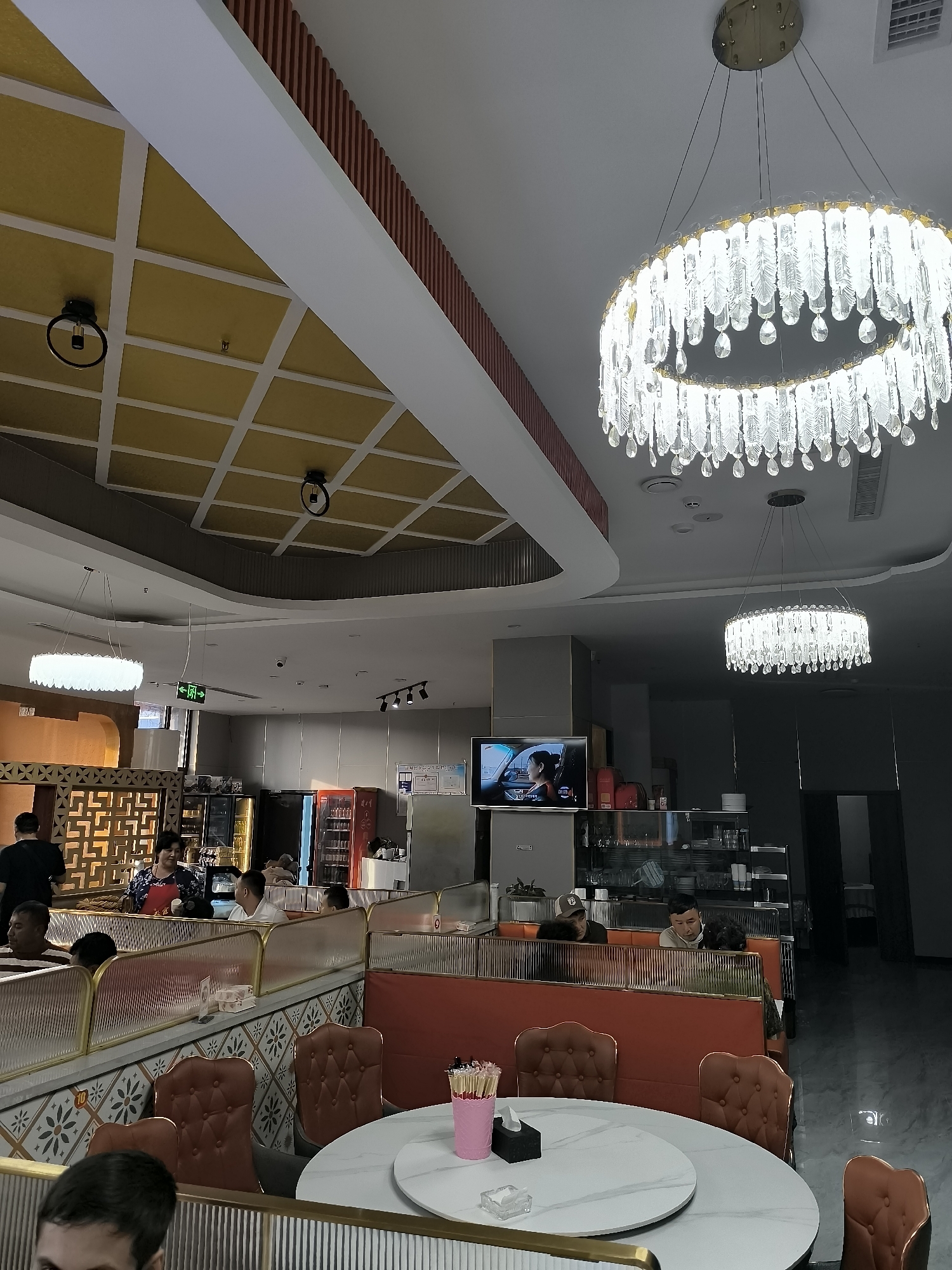 The image shows the interior of a restaurant, likely a hot pot restaurant, with patrons dining at tables. There are several decorative chandeliers hanging from the ceiling, and the seating appears to be in booths and around tables. The restaurant has a modern, somewhat upscale design with ornate ceiling tiles and patterned wall tiles. In the background, there are display cases with drinks and food items, and a television screen showing a scene with people. The restaurant is dimly lit, with bright chandeliers providing most of the illumination. The people in the image are mostly seated and engaged in eating or conversing. The overall impression is that of a busy dining establishment. This scene is located in Lucaogou Town, China.