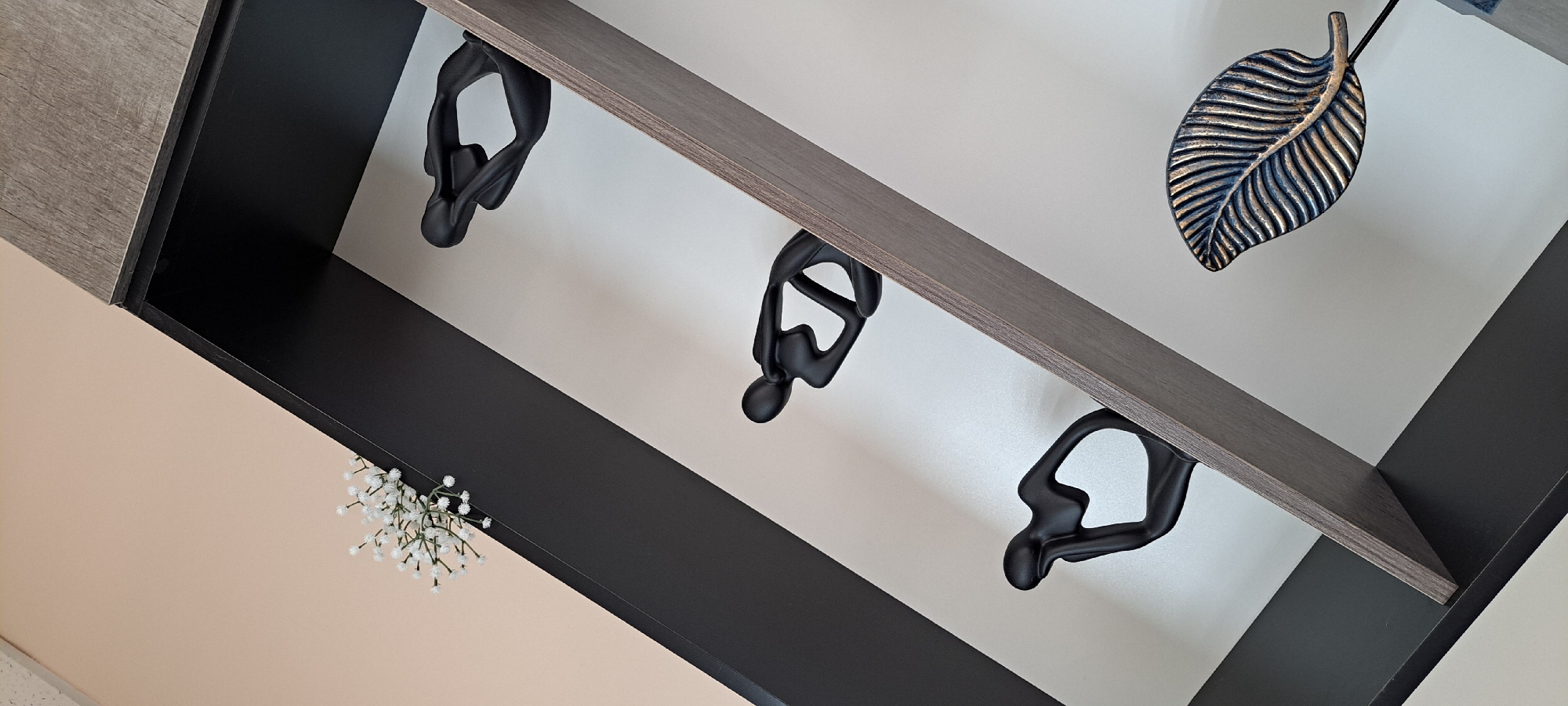 The image shows a black shelving unit with several decorative items. The shelves are angled and have a grey wood-grain trim. On the shelves, there are three black abstract sculptures of figures in a contemplative pose. There is also a white floral arrangement on the top left, and a metallic leaf sculpture on the bottom right. The background is a pale peach wall. There is no visible indication of the location being in Yessentuki, Russia.