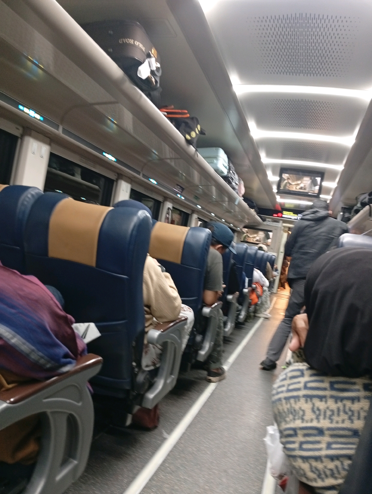 An interior view of a train carriage traveling through Kanci, Indonesia, shows rows of blue and tan seats with passengers seated or standing. Luggage is stored on overhead racks. A man in a black jacket and hood is walking down the aisle. The lighting is artificial, with overhead lights illuminating the carriage. The floor is gray and the windows on the side are visible.