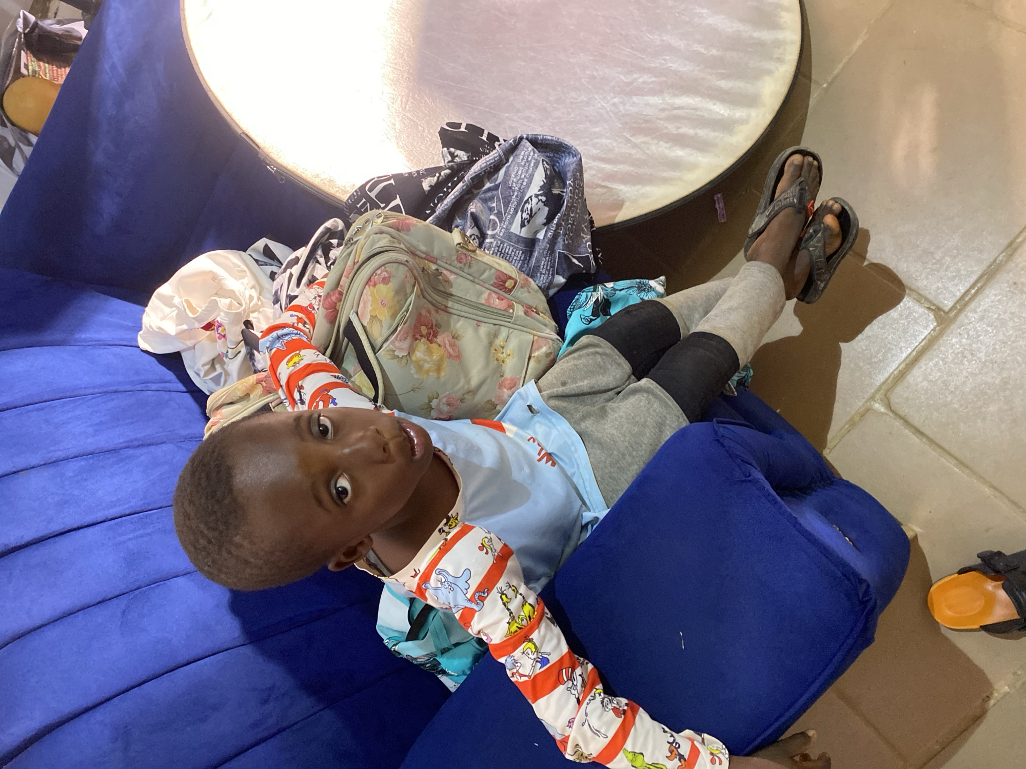 A young boy with short hair sits on a blue couch in what appears to be a living room in Barakin Akawo, Nigeria. He is wearing pajamas with a light blue top and sleeves decorated with red and white stripes featuring cartoon characters. His pants are gray. He is looking directly at the camera with his mouth slightly open. Next to him on the couch are several bags and folded clothing, including a floral-patterned bag and a bag with a newspaper-like print. A large, diffused light source is visible to the right of the boy, casting a soft glow on the scene. The floor is tiled.