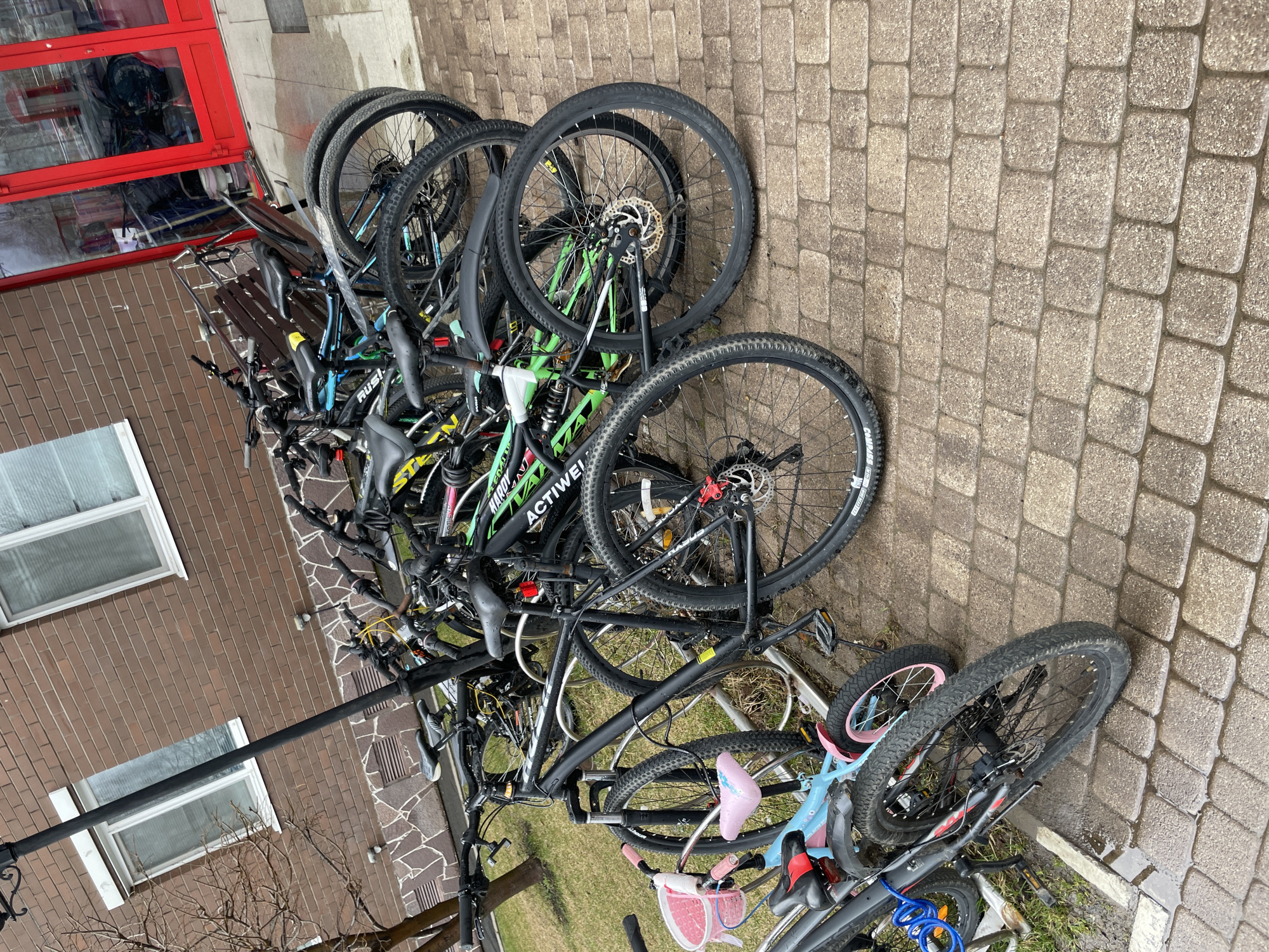 This image, captured in Izhevsk, Russia, presents a busy outdoor scene dominated by numerous bicycles parked against a brick building. A variety of bicycles, including several adult mountain bikes with disc brakes and knobby tires, are clustered together. They are either leaning against each other or secured to a simple metal bike rack. Among them is a small light blue child's bike with pink accents and a front basket.
The ground consists of rectangular paved stones, which appear damp or wet, suggesting recent rain or humid conditions. A small patch of grass is also visible on the right side of the frame, beneath some of the bikes.
To the left, a brown brick building facade is prominent, featuring white-framed windows with horizontal blinds or grates. A section of the building in the upper left corner has a distinctive red frame around what appears to be a large window or door. A dark, horizontal metal pole runs across the mid-ground, partially obscuring the building. Bare tree branches are visible in the lower left corner, indicating a non-foliated season.
Notable text visible on the bicycles includes "ACTIVWEI" on a green frame, "RUSI" on a dark-framed bike, "COMPASS" on a tire, and "STELL" on another bike frame. The lighting is overcast, consistent with a cloudy day, and there are no people present in the scene. The setting appears to be a practical, possibly communal, bike parking area outside an urban or suburban building.