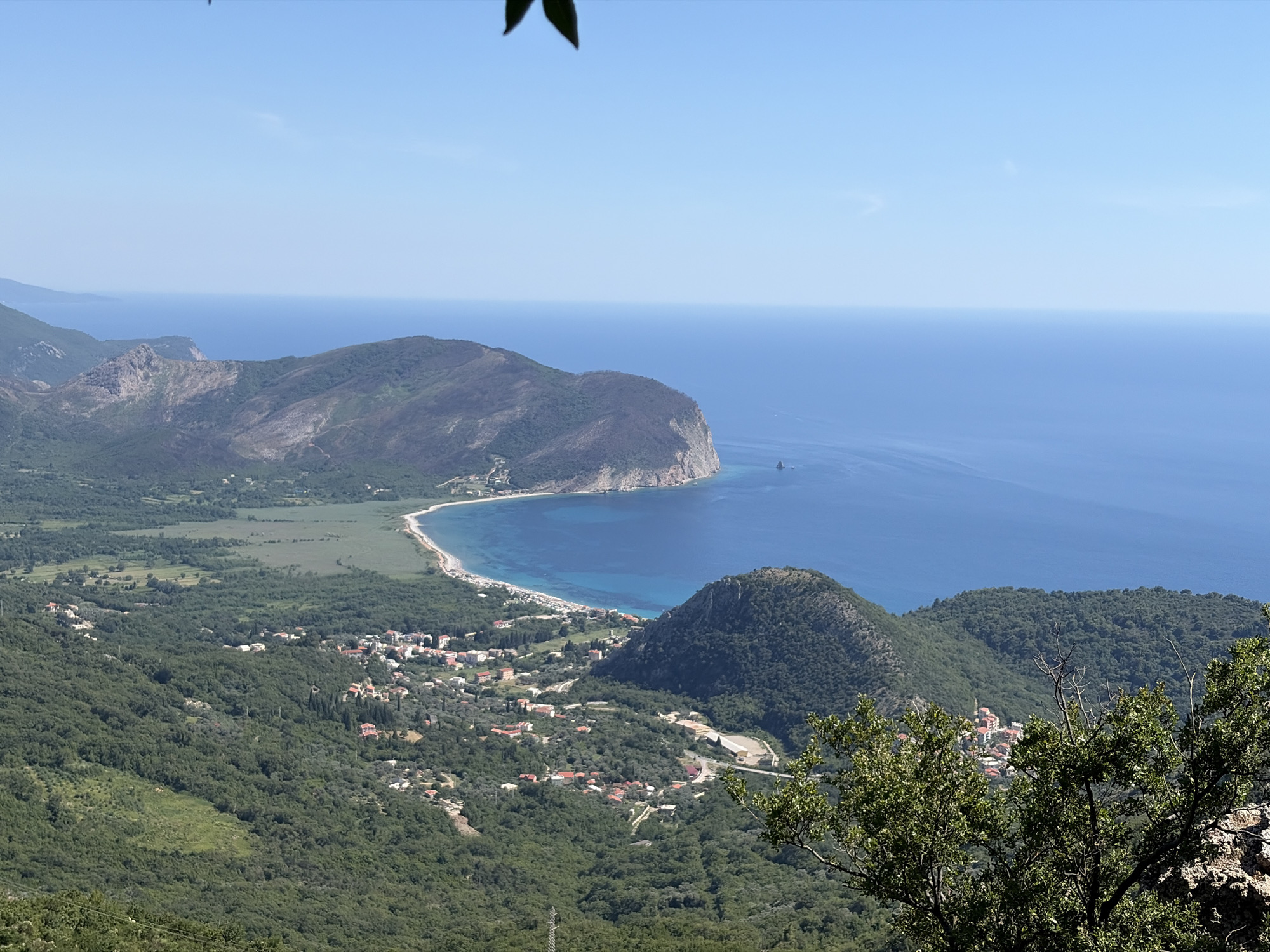 The image is an aerial view of the city of Novoselje, Montenegro, showcasing its coastal location. The scene is dominated by the vibrant blue Adriatic Sea meeting a curved coastline with a sandy beach dotted with umbrellas. Steep, verdant hills covered in dense, dark green trees rise from the coastal plain, interspersed with clusters of red-roofed buildings that form the town. A prominent, rounded, green hill is situated in the foreground, partially obscuring a view of a small town with red-tiled roofs nestled within the dense foliage. In the distance, a larger, rocky, mountainous terrain is visible, also covered in greenery, with a rugged, steep incline plunging towards the sea. The sky is a clear, bright blue, with the horizon line where the sea and sky meet being crisp and distinct. A single dark leaf is visible in the upper left corner of the frame, intruding slightly into the sky. The overall impression is one of a serene, sun-drenched Mediterranean landscape.