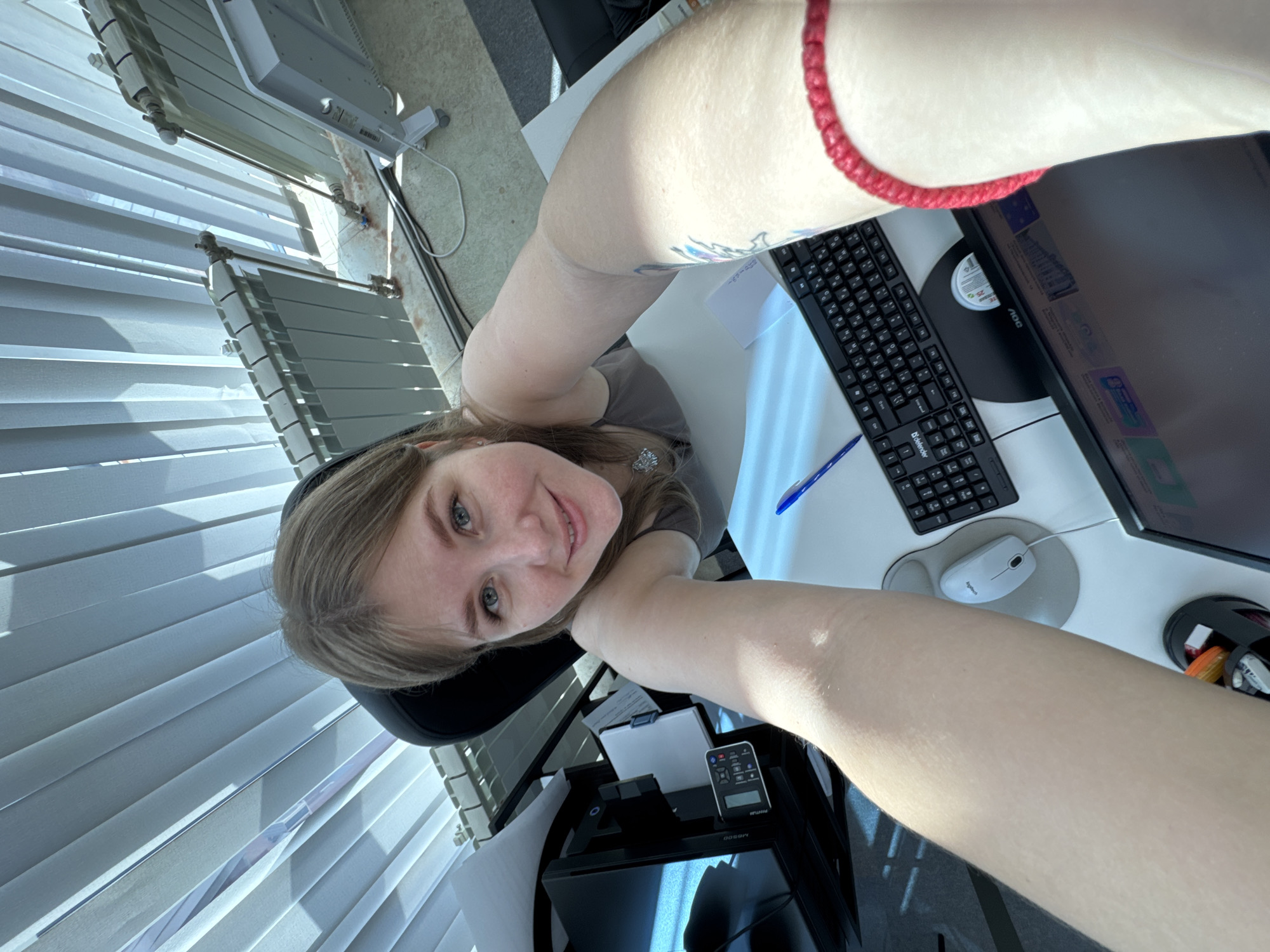 A person is seated at a desk in a room with vertical blinds on the window. The desk has a computer monitor, keyboard, mouse, pen, and printer. The person is looking up at the camera, taking a selfie. The location appears to be an office. The city of Perm, Russia, cannot be confirmed from the image.