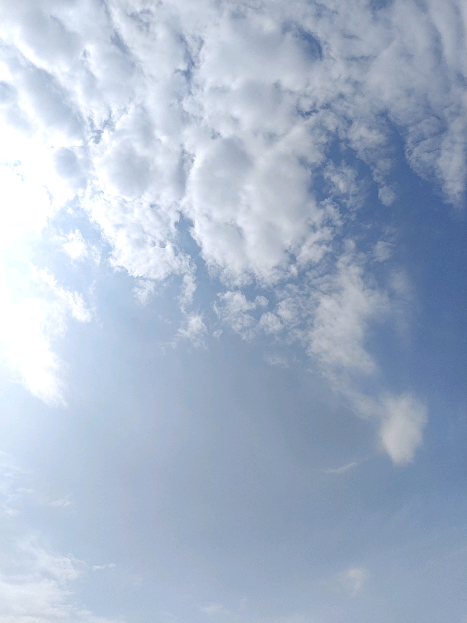 The image shows a sky with scattered clouds and a bright sun. The sky is a light blue, and the clouds are white and fluffy. The sun is partially obscured by the clouds. The image appears to have been taken on a clear day. There are no people, subjects, objects, or actions depicted in the image. The environment is purely sky and clouds. There is no indication of the city of Nellore, India, or any other specific location.