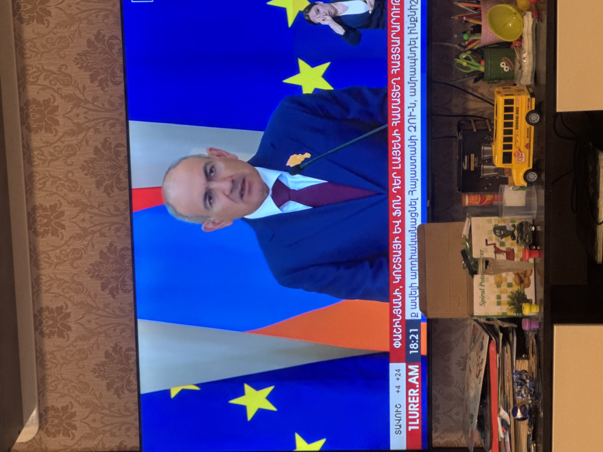 The image shows a television screen displaying a news broadcast in Yerevan, Armenia. The screen features a balding man in a blue suit and red tie, addressing the audience in front of a backdrop that includes elements of the EU flag. A smaller inset image shows a woman in a black suit and white shirt. The broadcast's text mentions "ՓԱՇԻՆՅԱՆԻ, ԿՈՇՏԱՅԻ" and "LURER.AM 18:21".
Surrounding the television are various household items, including a toy school bus, a green container with a face design, pencils, scissors, a yellow bowl, a "Spiral Potato" box, and several books and papers.