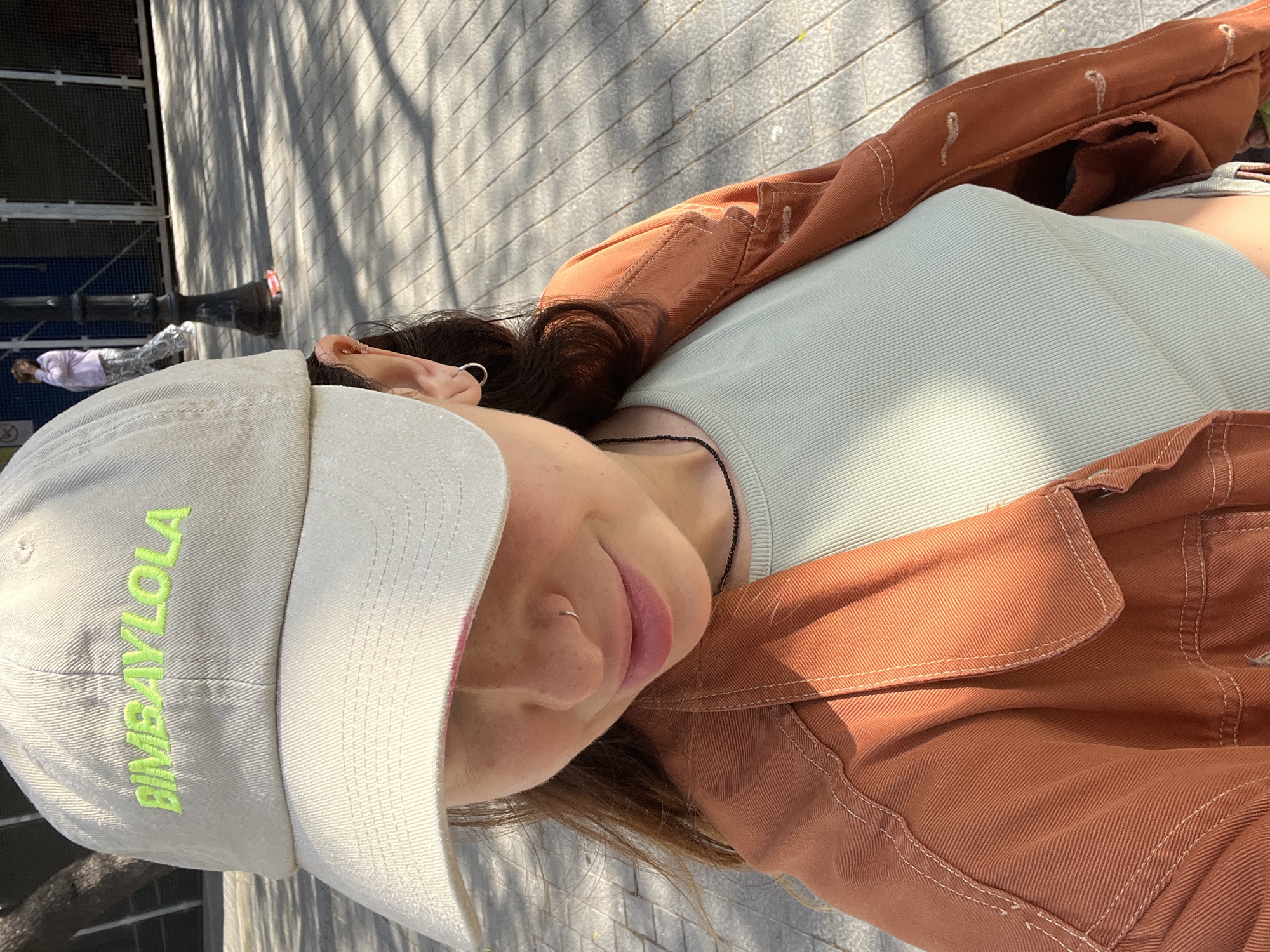 A close-up, slightly Dutch-angled shot captures a young person from the chest up, positioned outdoors in what appears to be an urban setting, likely Barcelona, Spain, given the location context. The person is looking directly at the camera with a slight smile, showing a relaxed expression.
They are wearing a light-colored, possibly off-white, baseball cap with "BIMBA Y LOLA" embroidered in neon green text across the front. Dark brown hair spills out from under the cap, framing the face. Visible facial features include light freckles, soft pink lips, and a small silver hoop piercing in the nose. Multiple earrings are seen on the left ear: a small gold stud/hoop on the helix and a larger silver hoop on the lobe.
The individual is dressed in a light mint green ribbed tank top, layered under a rust-colored or burnt orange jacket made of sturdy fabric, possibly denim or canvas. The jacket features prominent white stitching along the seams and pockets. A thin, black beaded necklace is visible around the neck, just above the tank top.
The background shows a paved surface, likely a sidewalk or plaza, with strong shadows indicating bright sunlight. In the distance, other faint figures can be seen, along with a dark lamppost and a metallic grid-like structure, possibly part of a building or fence. The overall impression is a casual, sunny day in a city environment.