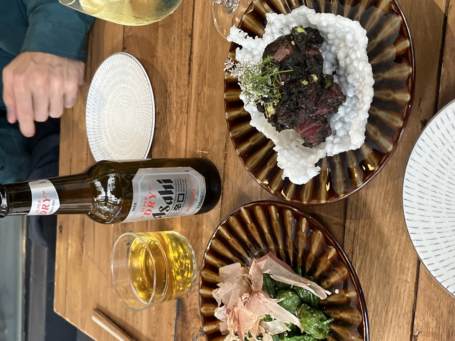 The image displays a dining table scene within a restaurant in Mexico City, Mexico. A light brown, rustic wooden table serves as the primary surface. At least one individual is present, with hands and forearms visible. Central to the composition is a partially consumed 330ml bottle of Asahi Super Dry beer. To its left, a clear glass holds an amber liquid, approximately half full. To the right, a stemmed glass contains a pale yellow liquid. Two brown, ribbed ceramic plates are in the foreground. The left plate contains green peppers topped with light brown, shaved flakes. The right plate holds a dark, red-hued, cubed food item resting on an irregular white crispy base, garnished with small white flowers and green foliage. An empty white plate with a radial pattern is positioned behind the beer bottle, and a similar plate is partially visible in the lower foreground. A pair of light-colored chopsticks rests on the table to the far left. The background is blurred, indicating an indoor dining environment. The scene depicts a meal involving Japanese-style dishes and beverages.